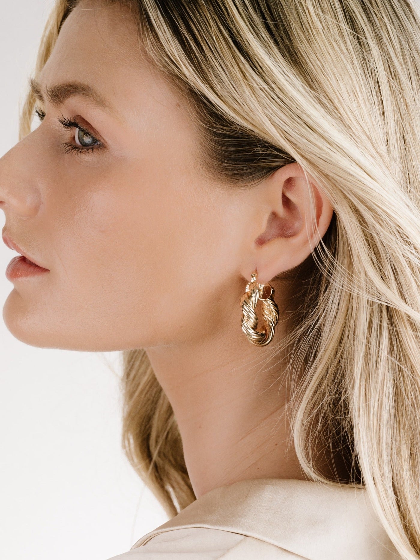 A woman with long, wavy blonde hair is wearing stylish golden drop earrings. She is facing slightly to the side against a light background, highlighting her facial profile and accessories.