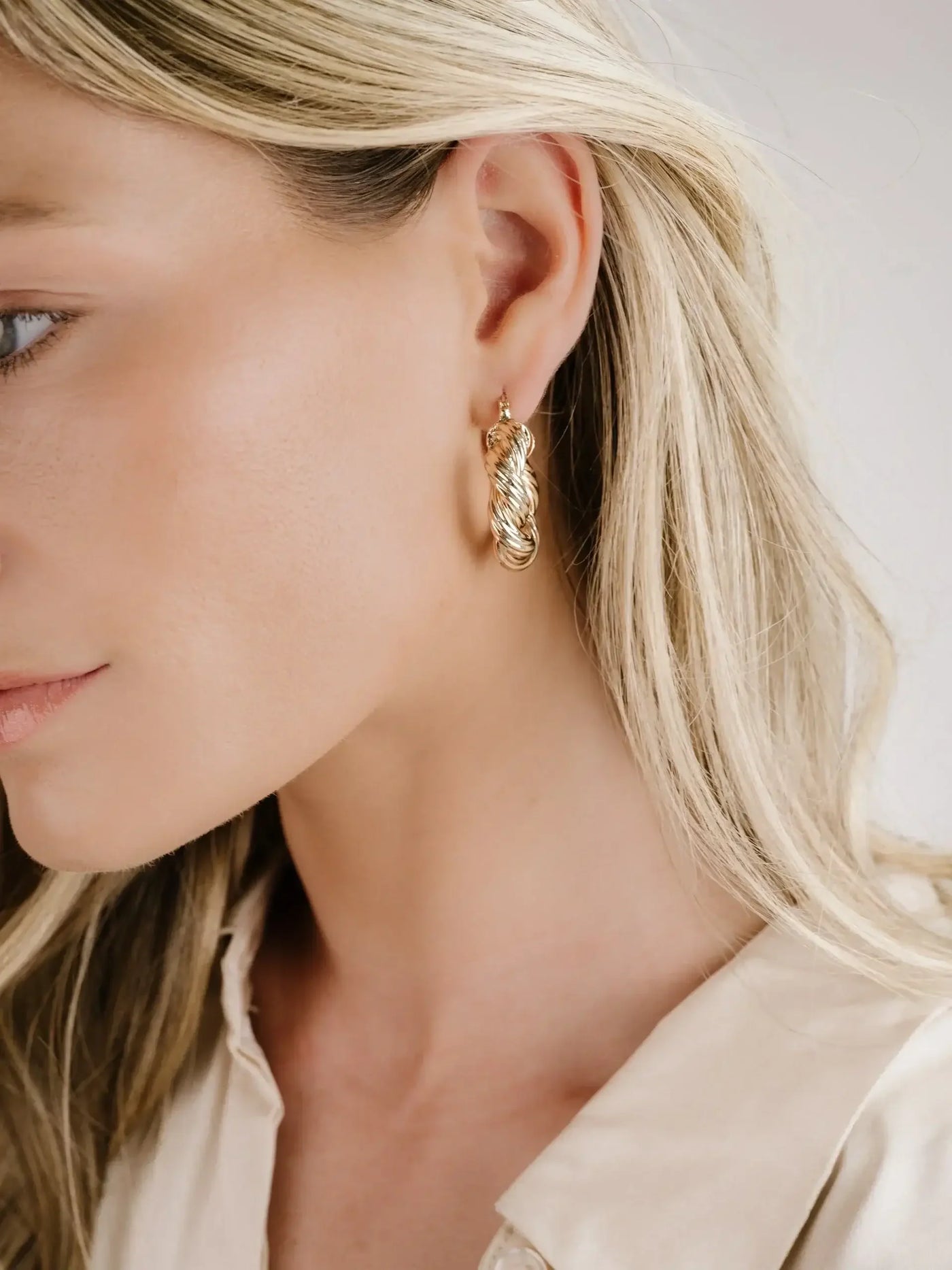 A close-up view of a woman's profile, showcasing a braided gold hoop earring on her ear. She has long, straight blonde hair and touches of natural makeup, set against a neutral background.