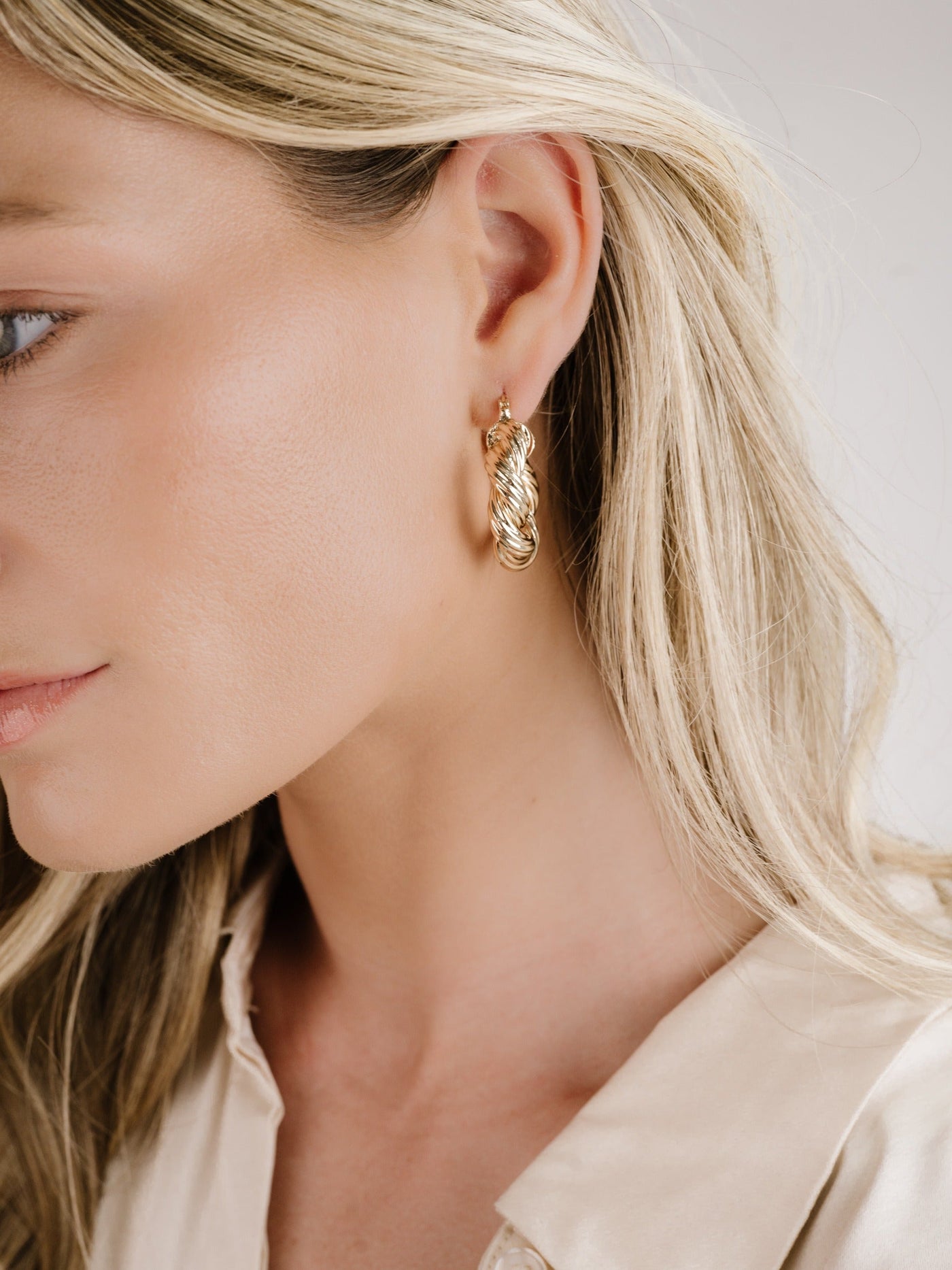 A close-up view of a woman's profile, showcasing a braided gold hoop earring on her ear. She has long, straight blonde hair and touches of natural makeup, set against a neutral background.