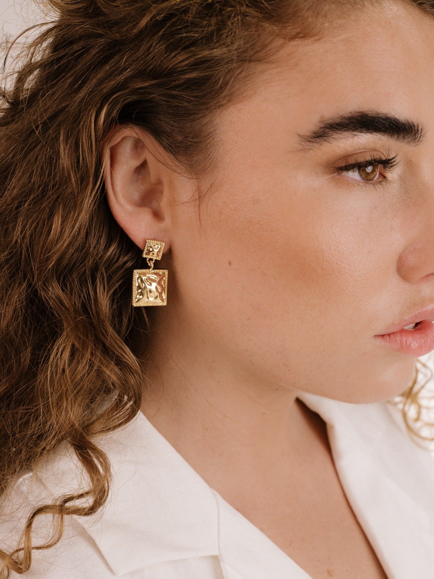 A woman with curly hair wears stylish gold earrings featuring square designs. The earrings hang closely to her face in a soft, neutral-toned setting, highlighting her elegant features.