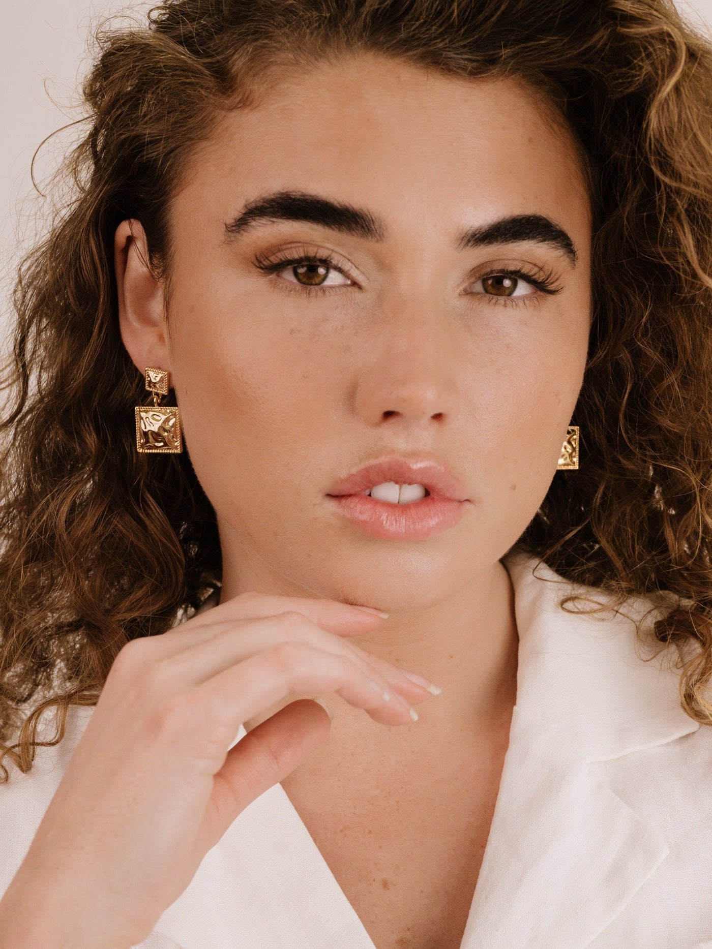 A young woman with curly hair poses closely, lightly touching her chin. She wears bold gold earrings and has natural makeup against a neutral background, highlighting her facial features.