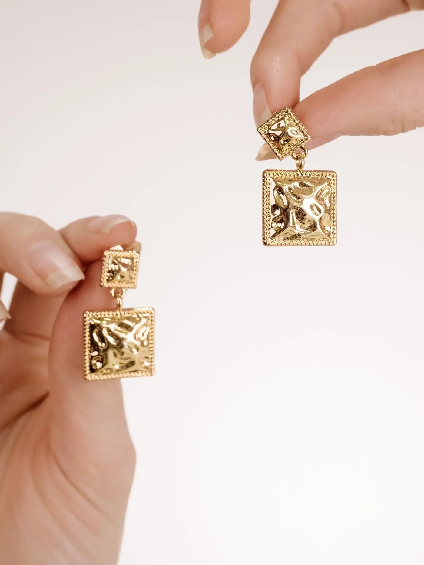 Two pairs of gold earrings are held up by fingers. The earrings feature a textured design with a square shape. The background is a soft, neutral color.