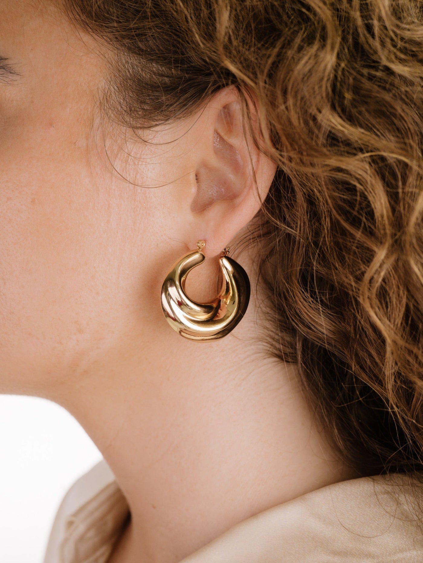 A large, shiny gold hoop earring is worn on a woman’s ear, complementing her wavy hair and light skin tone, set against a soft, neutral background.