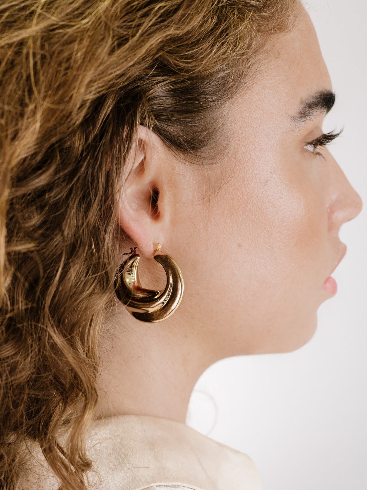 A woman with curly hair wears a large, glossy gold hoop earring. She gazes slightly off to the side, with a soft, muted background highlighting her style.