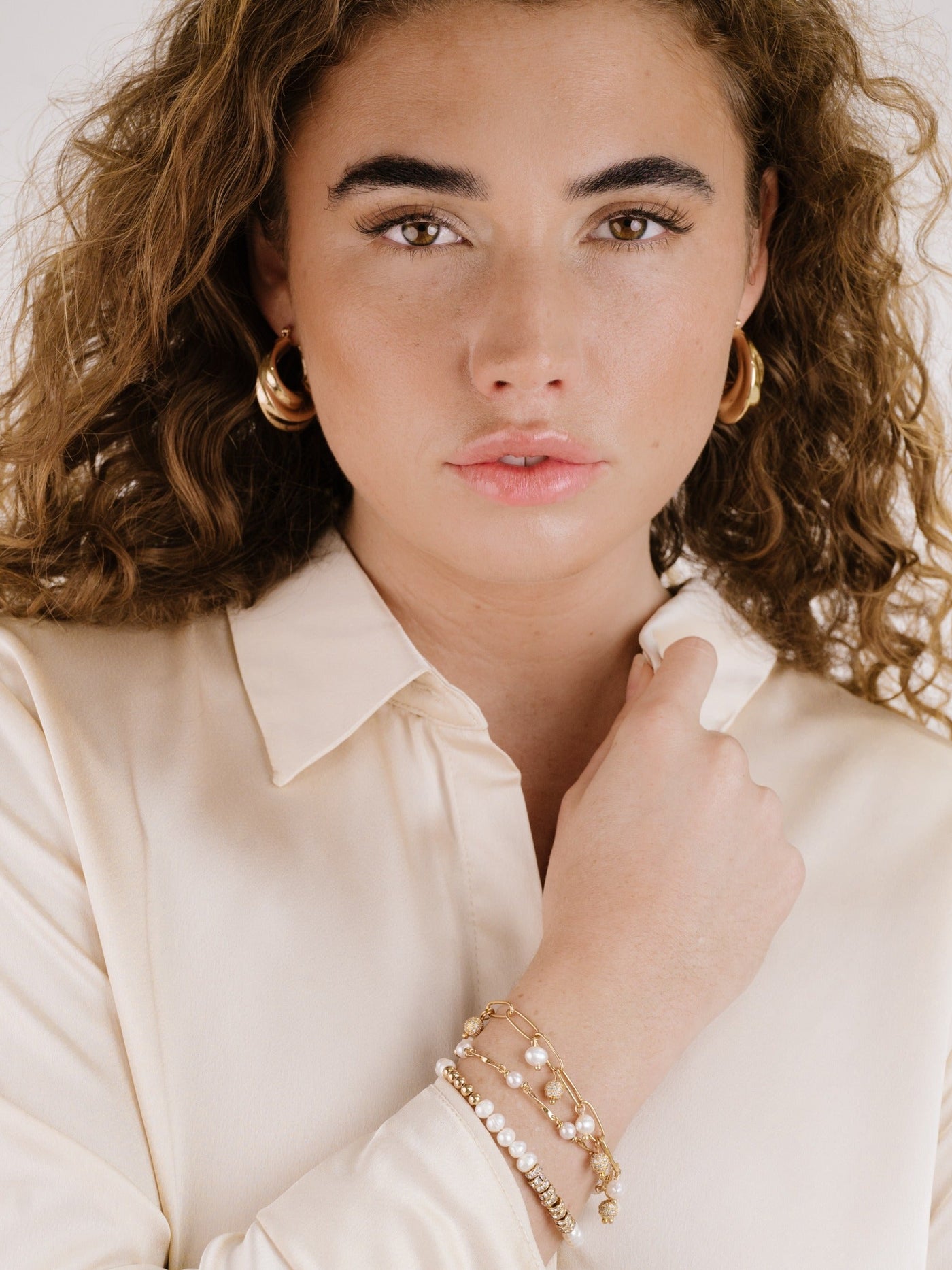 A young woman with curly hair poses, wearing a cream-colored blouse and gold hoop earrings. She gently touches her neck with one hand, showcasing multiple bracelets on her wrist.