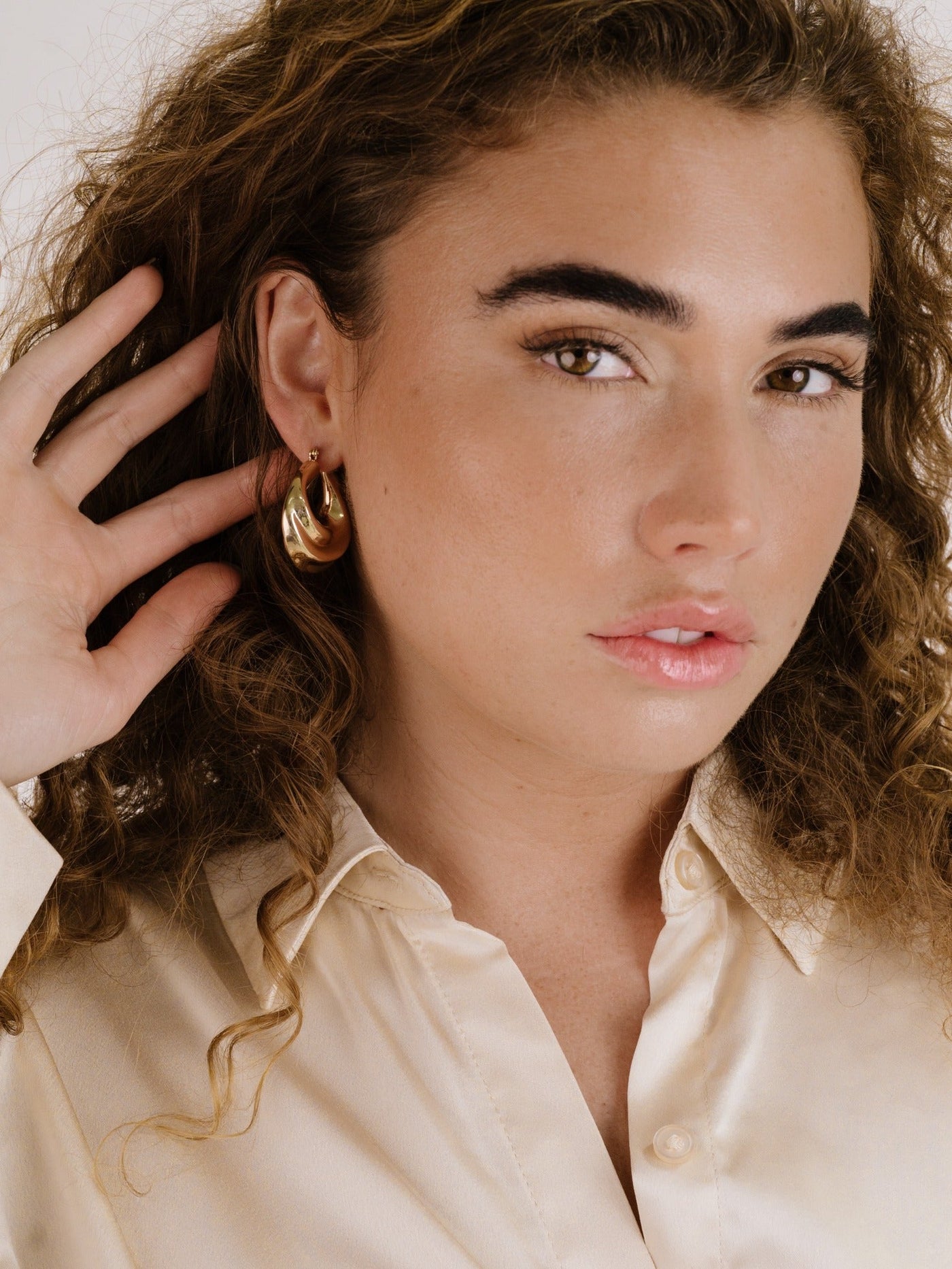 A young woman with curly hair holds her ear, showcasing a large, shiny gold earring. She wears a light, collared, satin shirt and gazes directly at the camera, exuding a confident expression.