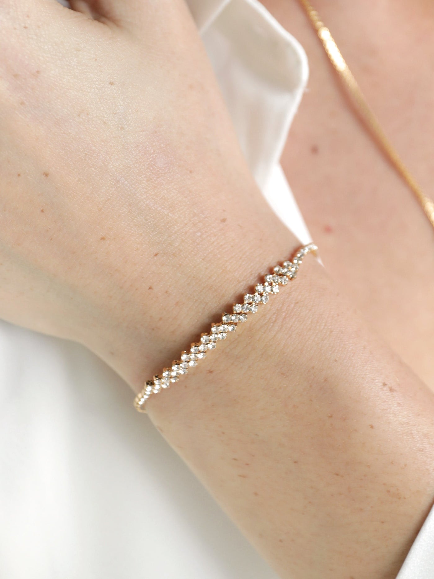A delicate gold bracelet adorned with small sparkling stones is worn on a person's wrist. The hand is partially visible against a soft, light-colored fabric background.
