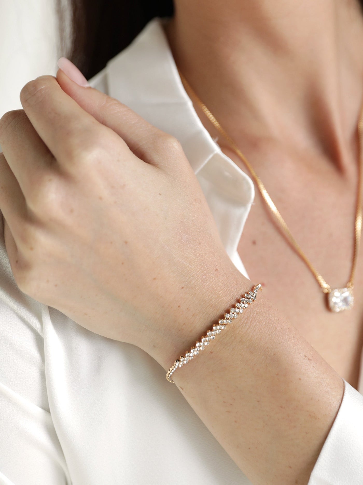 A delicate, sparkling bracelet adorned with small gems glimmers on a hand resting against a white shirt. A subtle gold necklace adds to the elegant appearance in a soft, neutral setting.