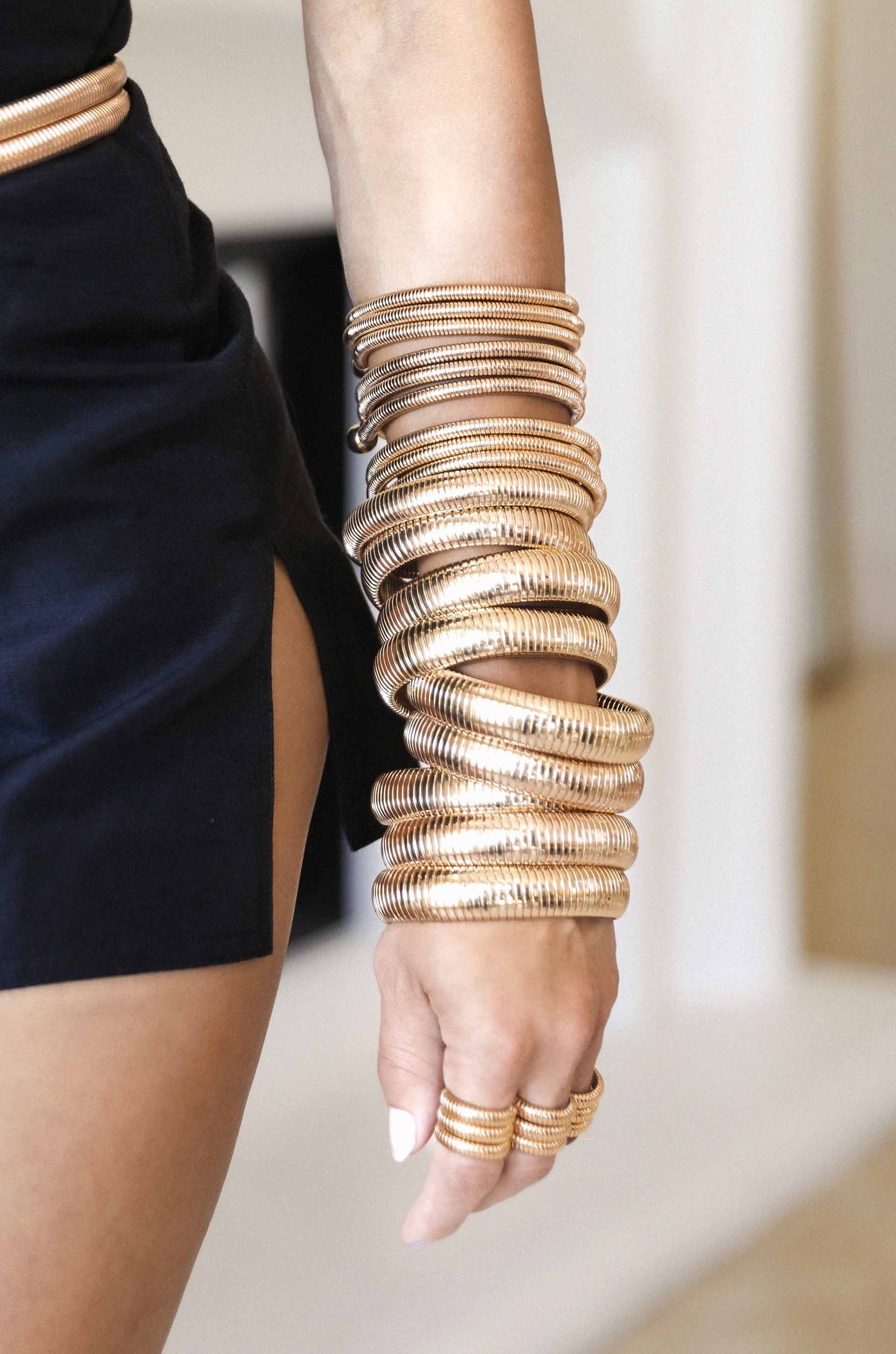 A slender arm adorned with multiple shiny, spiral bangles reflects light, while wearing a black outfit with a high slit. The background appears softly blurred, suggesting an indoor setting.
