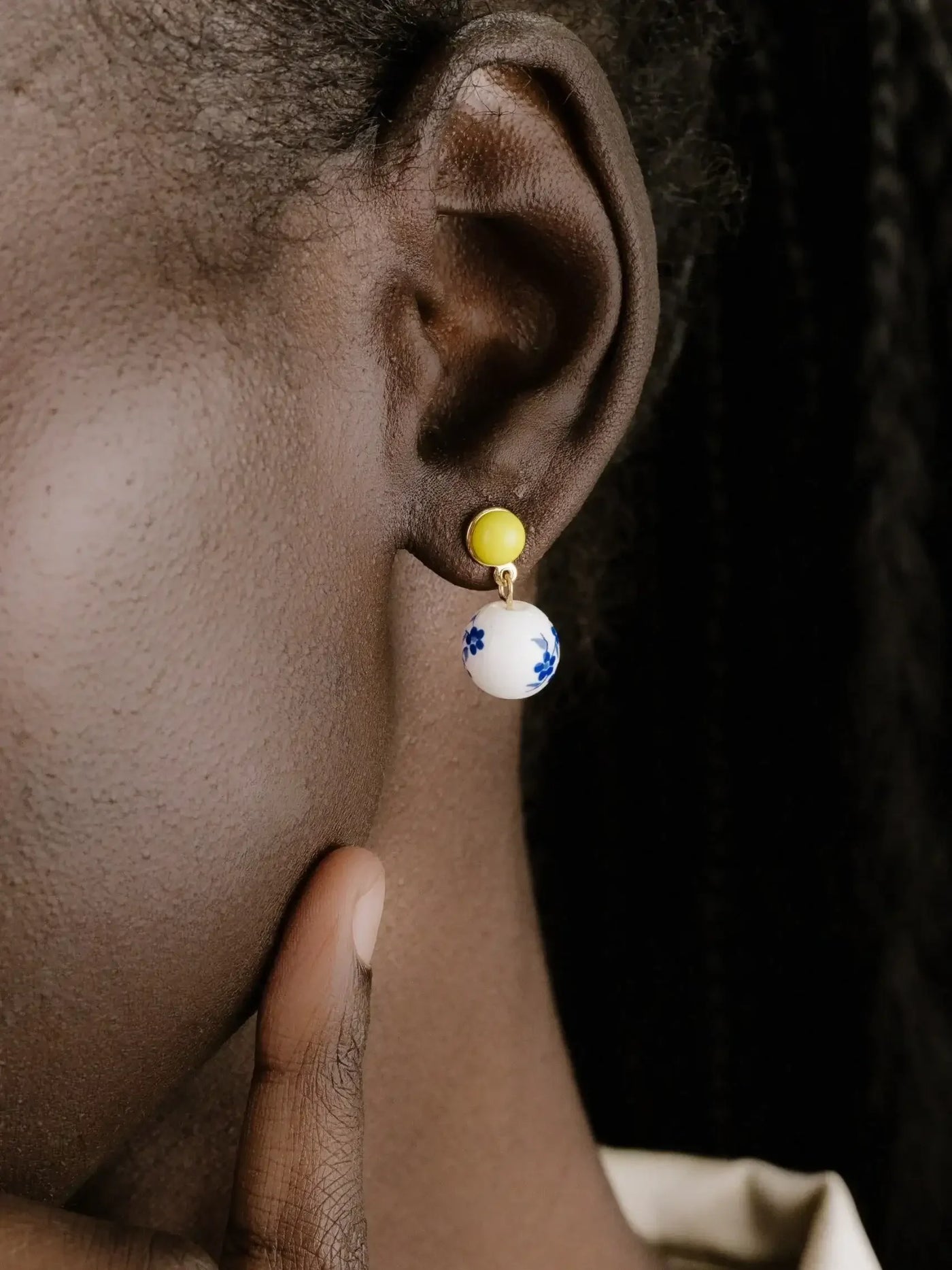 A close-up of an ear featuring a colorful earring with a yellow bead and a white sphere decorated with blue flowers. A hand gently touches the skin below the ear.