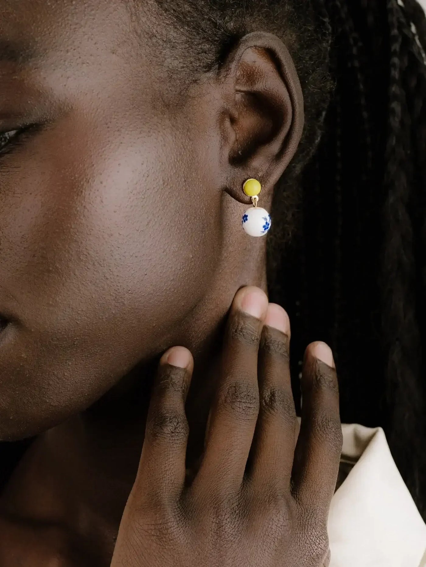 A person gently touches their neck while showcasing colorful earrings: a small yellow stud and a larger white ball adorned with blue floral designs, set against a plain background.