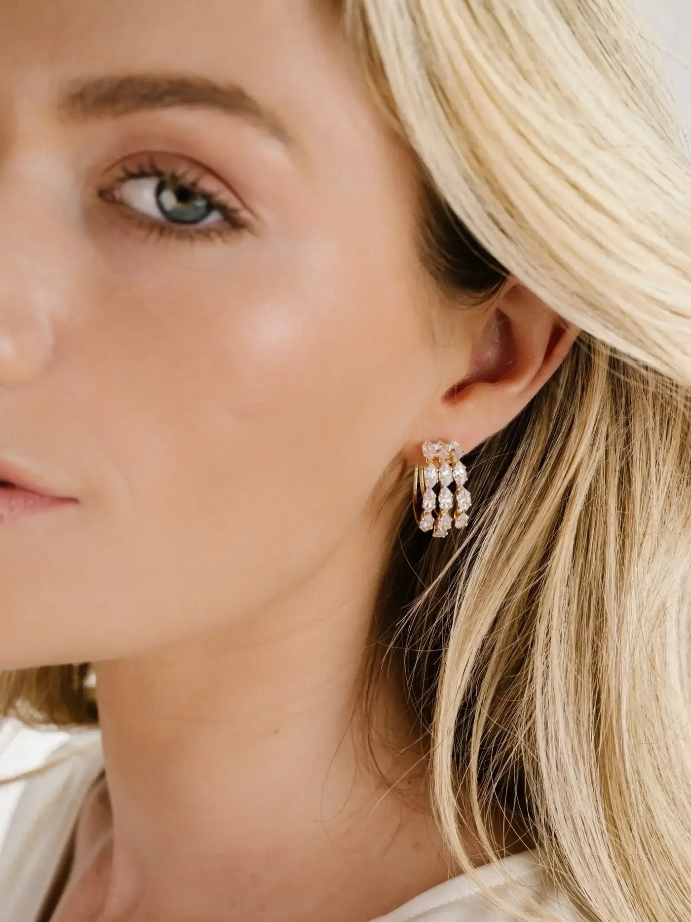 A young woman with light blonde hair wears elegant, sparkling earrings. She gazes to the side, with soft lighting highlighting her facial features and the jewelry.