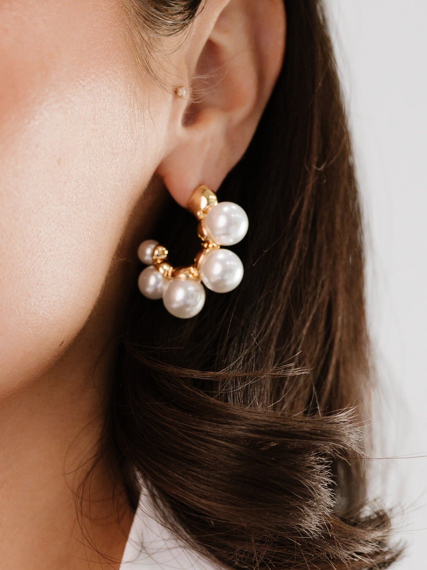 A close-up of a woman's ear showcasing a chunky gold earring adorned with multiple large white pearls, set against a soft, neutral background, emphasizing the elegant accessory.