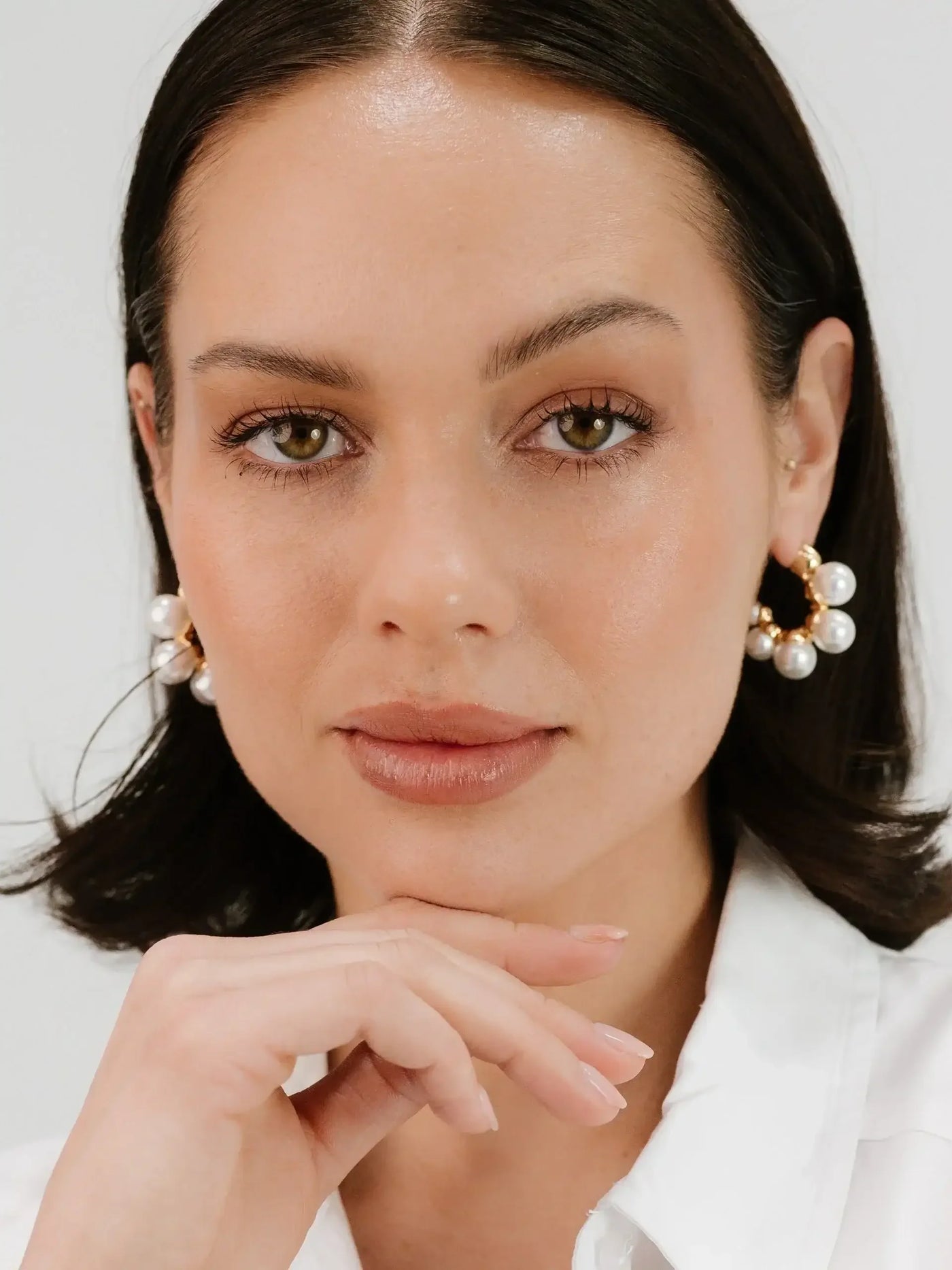 A woman with medium-length dark hair poses with a thoughtful expression, resting her chin on her hand. She wears large pearl earrings and a white shirt, set against a light background.