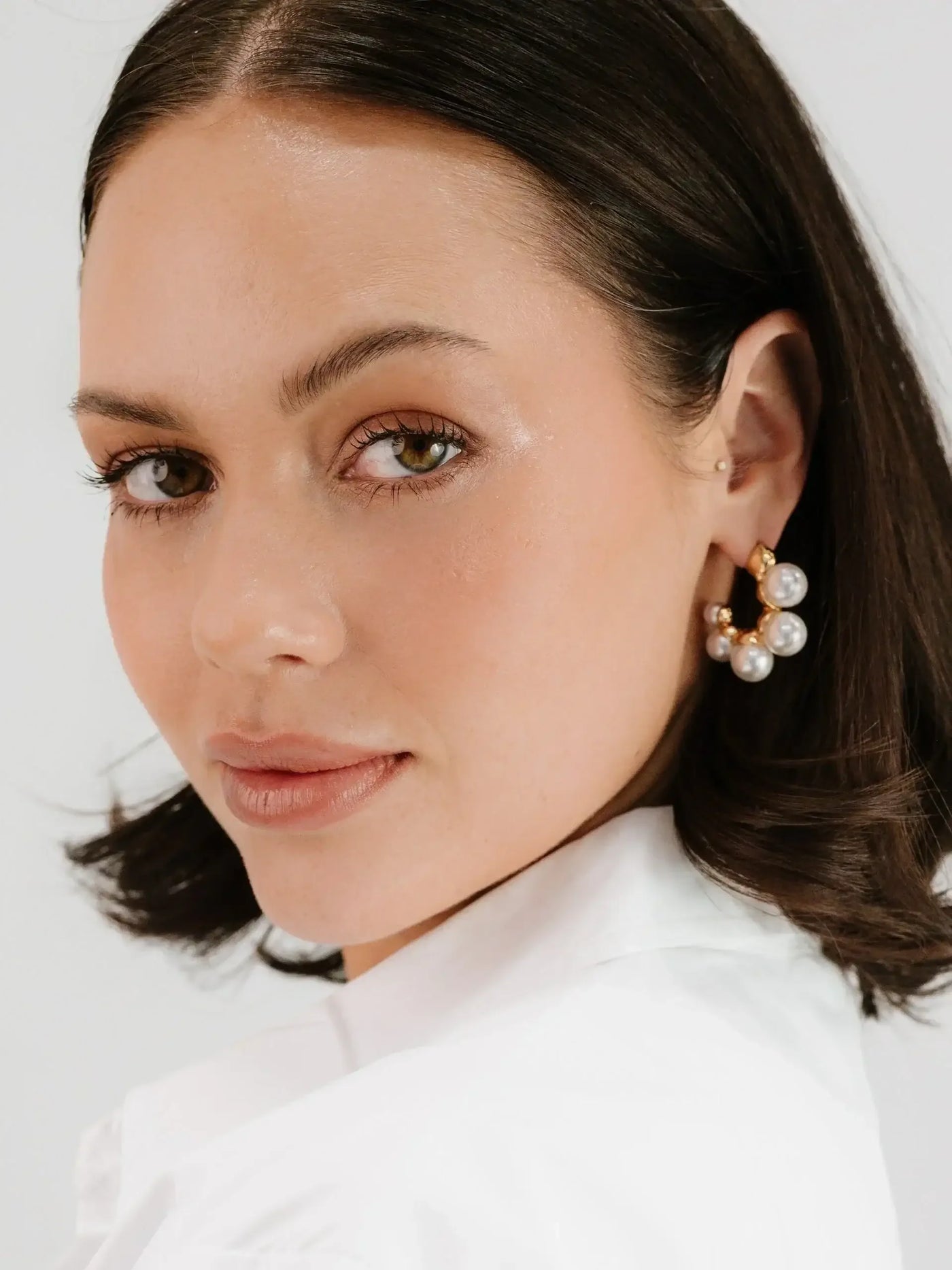 A woman with medium-length dark hair is wearing bold pearl earrings. She is slightly turned, gazing towards the viewer, and is dressed in a white top against a light background.