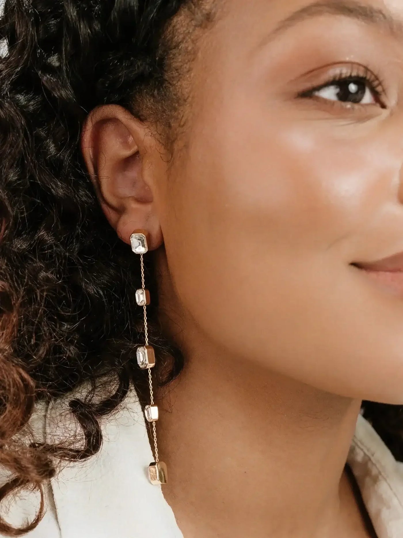 A close-up of a person’s ear adorned with a long, delicate earring featuring multiple geometric shapes, set against a soft, neutral background that enhances the earring's shine.