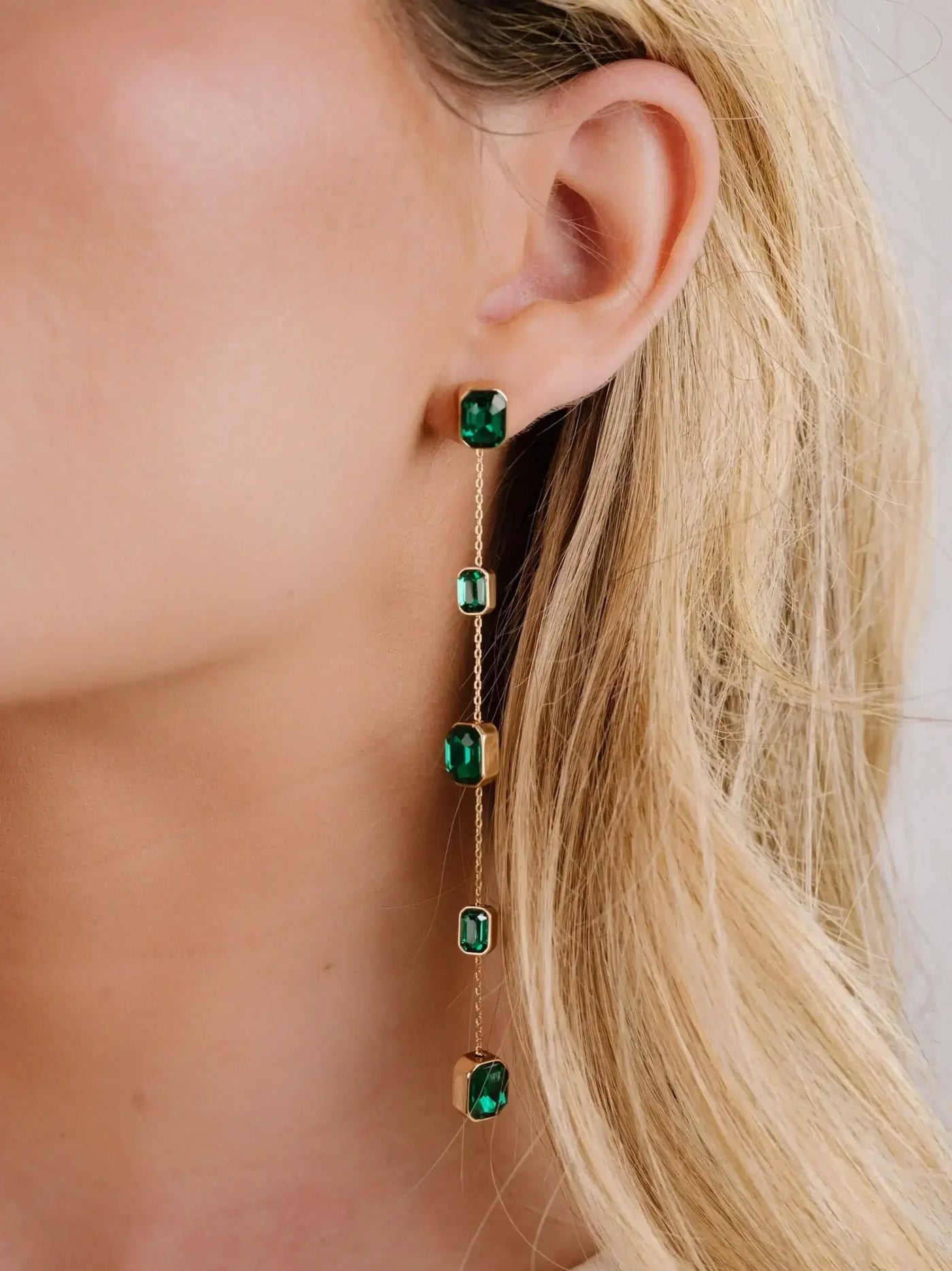 A long, elegant earring with green gemstones dangles from a woman’s ear, showcasing intricate gold settings against her light skin and flowing blonde hair, captured in a close-up view.