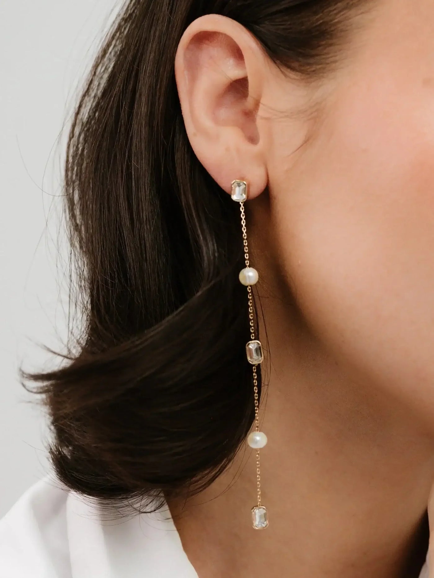 A dangling earring featuring a gold chain with alternating clear gemstones and a pearl is worn on a woman’s ear. Her dark hair softly frames her face.