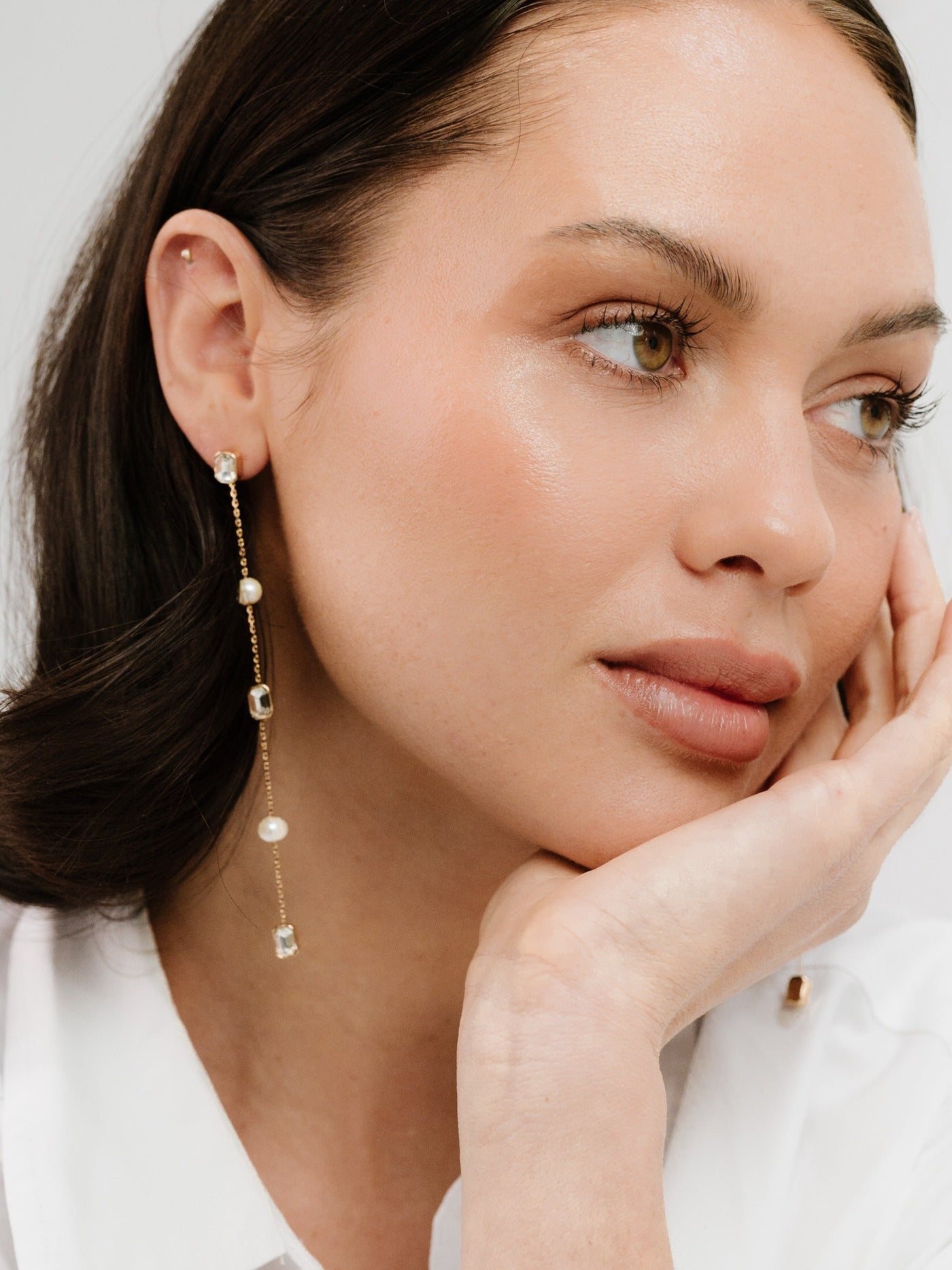 A woman with a natural makeup look rests her chin on her hand, wearing elegant, dangling earrings with pearls and gemstones against a soft, light-colored background.