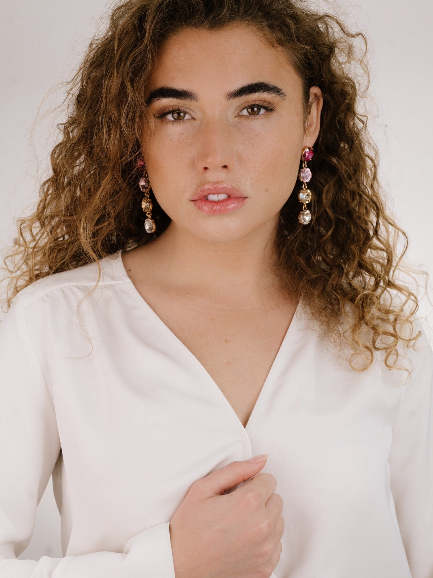 A woman with curly hair wears a white blouse and sparkling earrings. She gazes directly at the camera, with one hand gently touching her shirt, in a minimalistic studio setting.