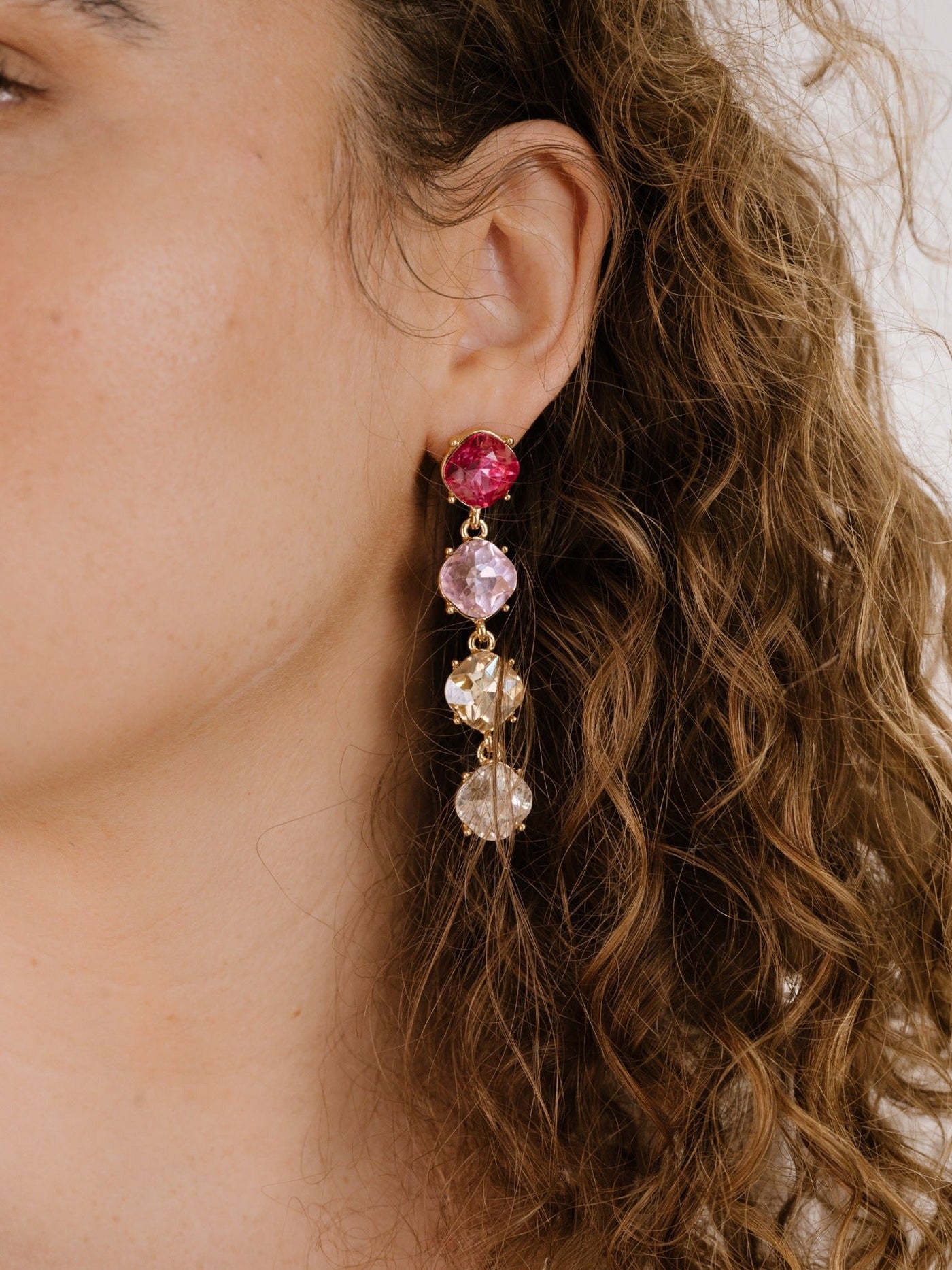 A large, multi-colored earring featuring pink and clear gemstones hangs from a person's ear, accentuating their curly hair and natural skin tone in a neutral setting.