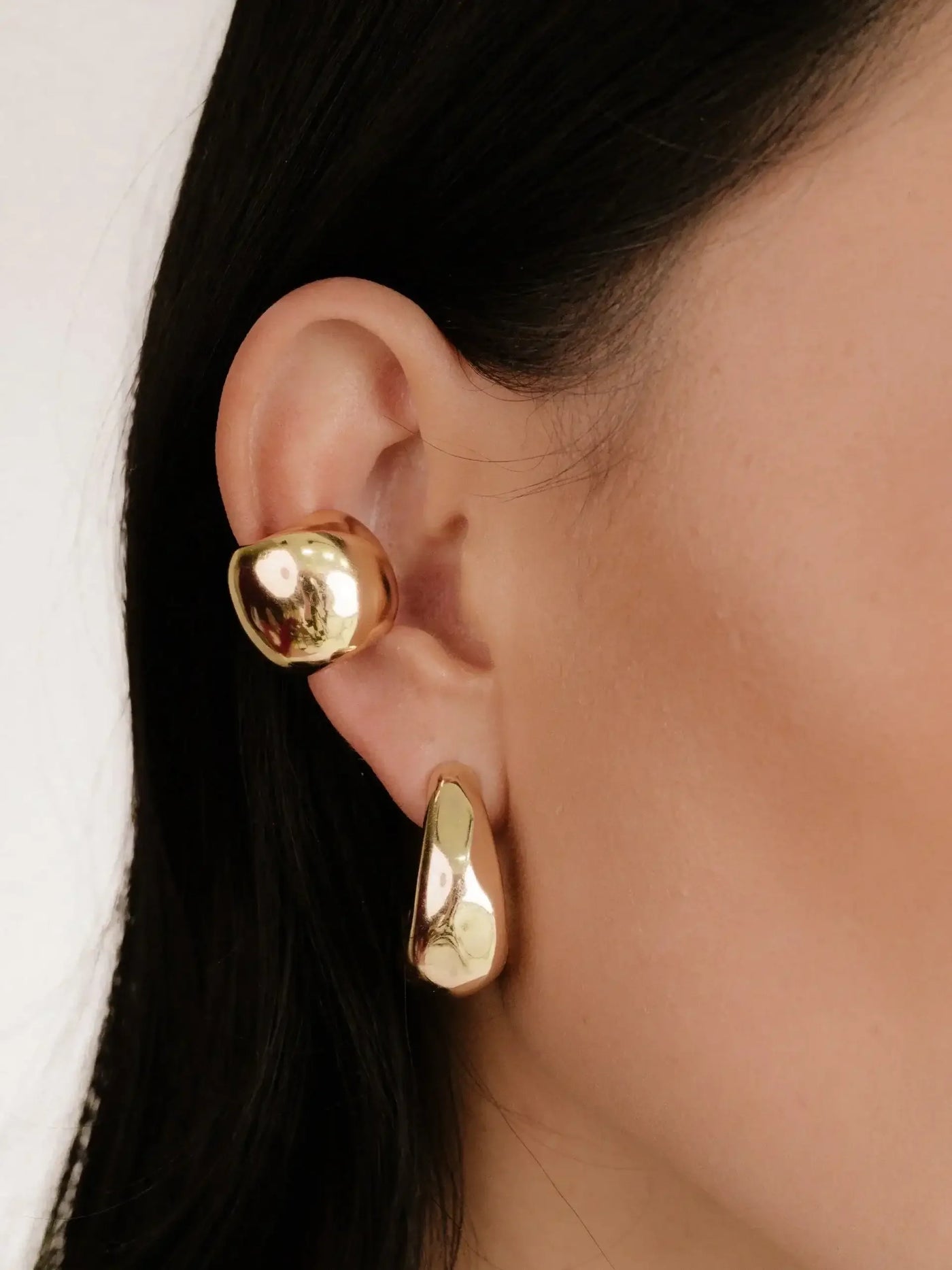 Two golden earrings are worn on a woman's ear. The earrings feature distinct, abstract shapes, catching light and reflecting a shiny surface. The background is neutral, highlighting the jewelry.