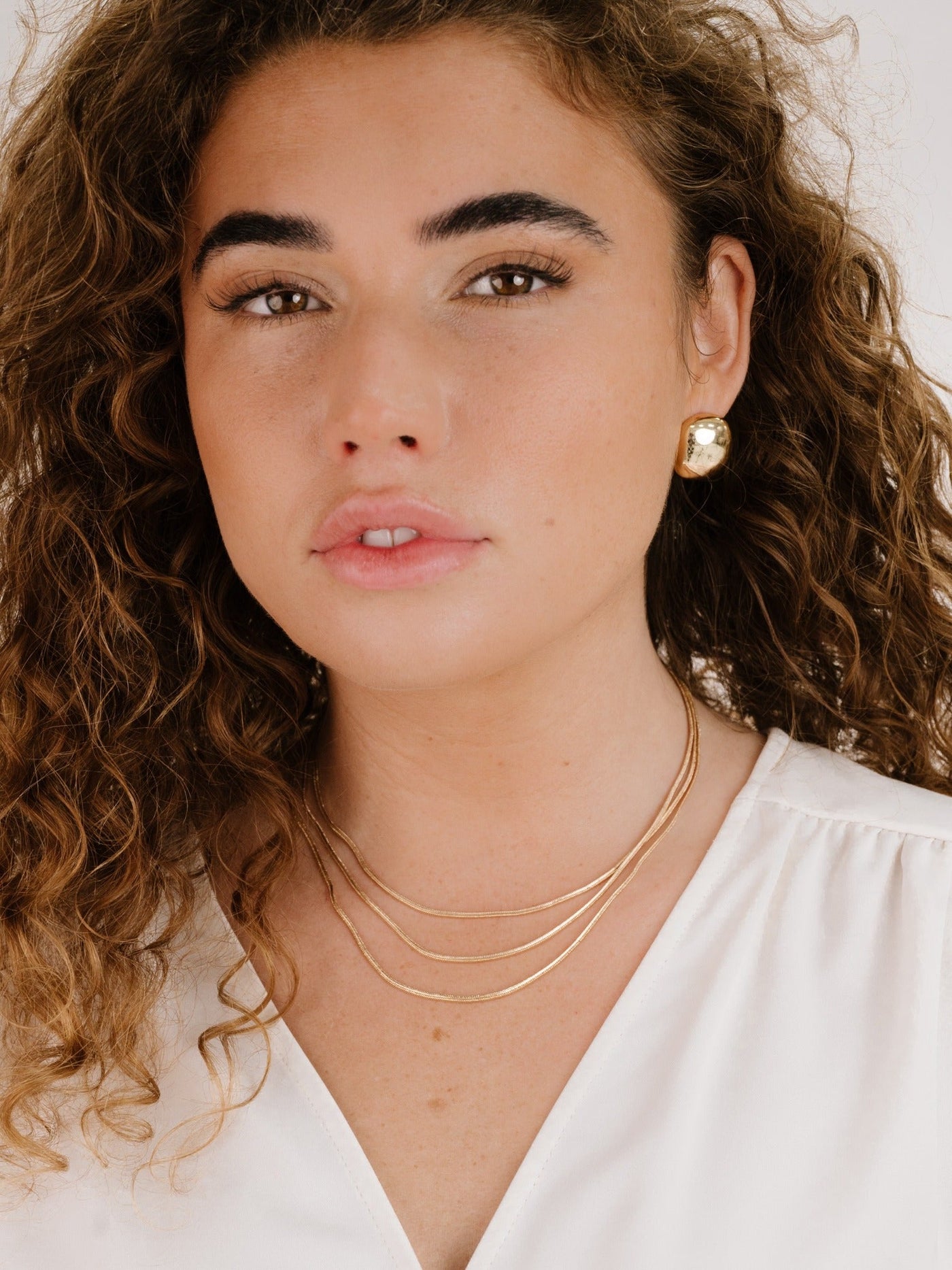 A person with curly hair and defined eyebrows gazes forward. They wear a white blouse, multiple gold necklaces, and a large gold earring, set against a neutral background.