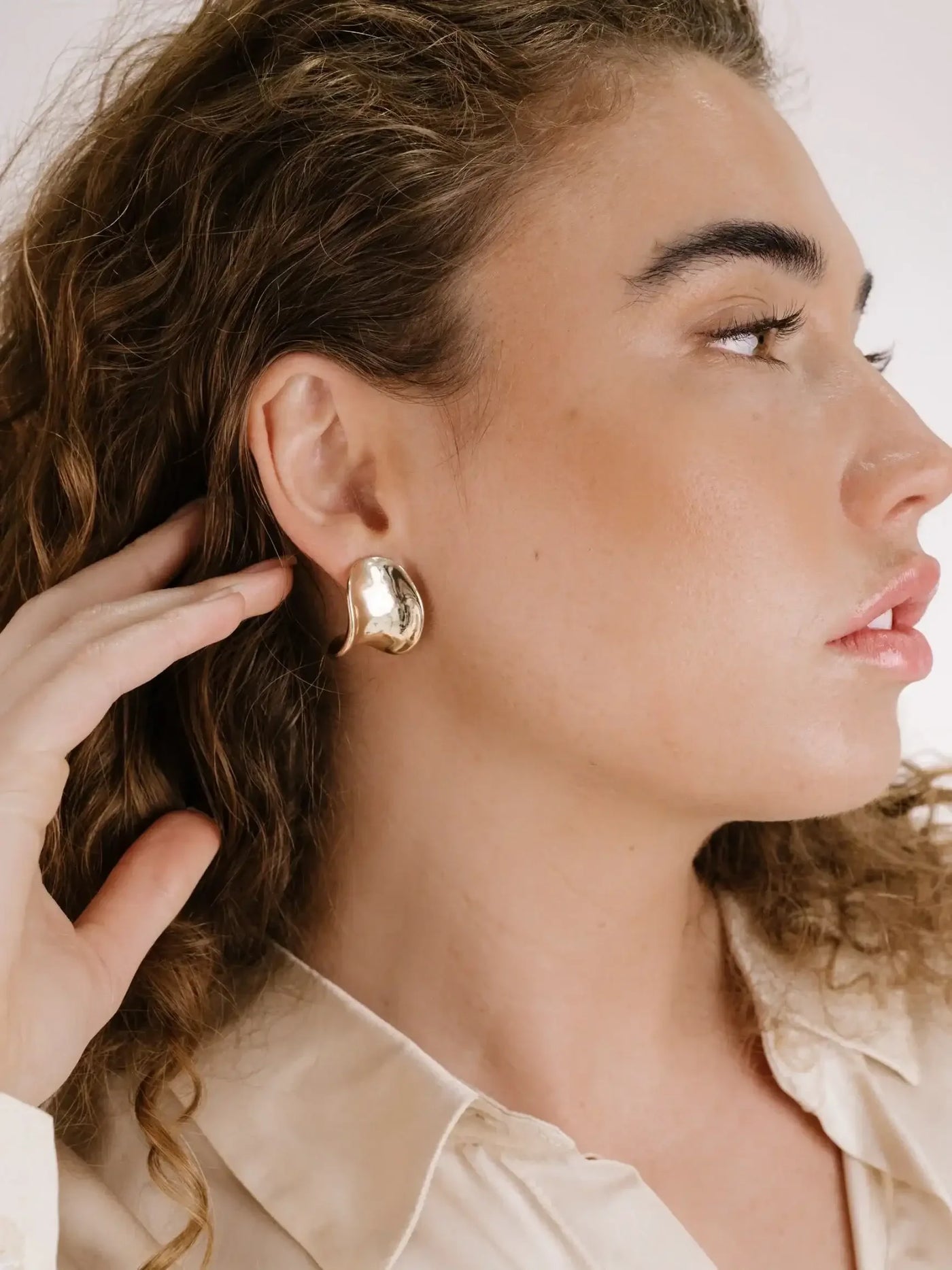 A woman with curly hair is touching a large, shiny gold earring on her ear, showcasing its design while wearing a beige button-up shirt against a neutral background.