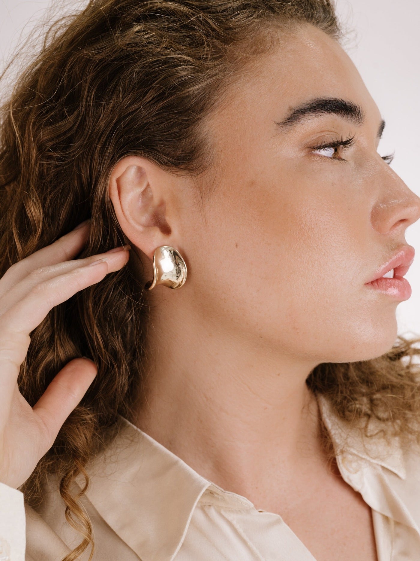 A woman with curly hair is touching a large, shiny gold earring on her ear, showcasing its design while wearing a beige button-up shirt against a neutral background.