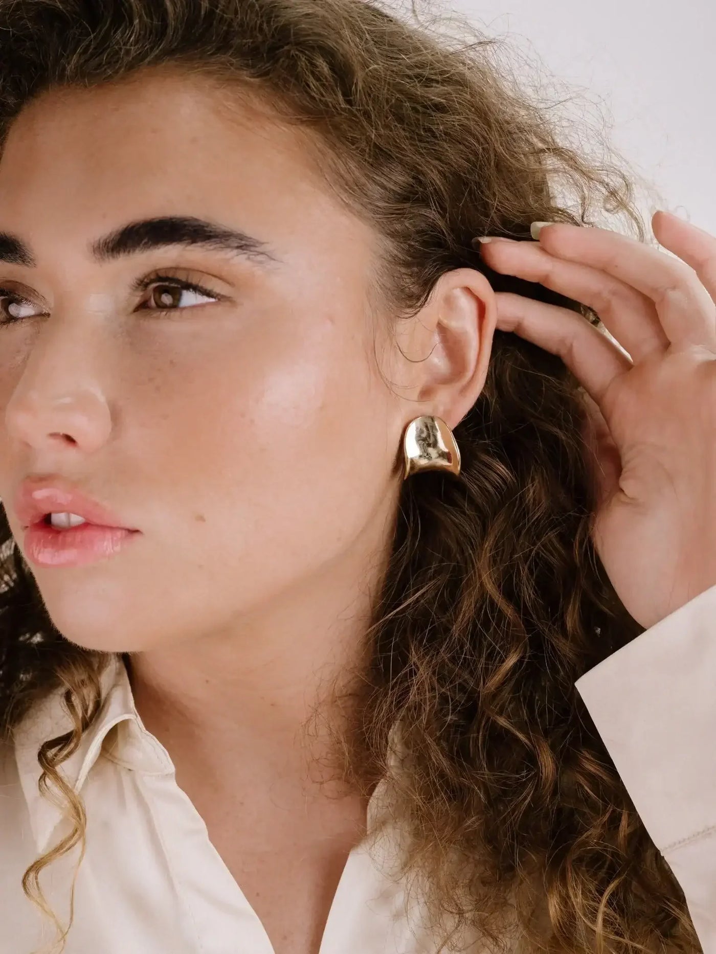 A close-up of a woman with curly hair touching her earlobe, showcasing a large gold earring. She is wearing a light-colored shirt, and her expression is calm and contemplative.