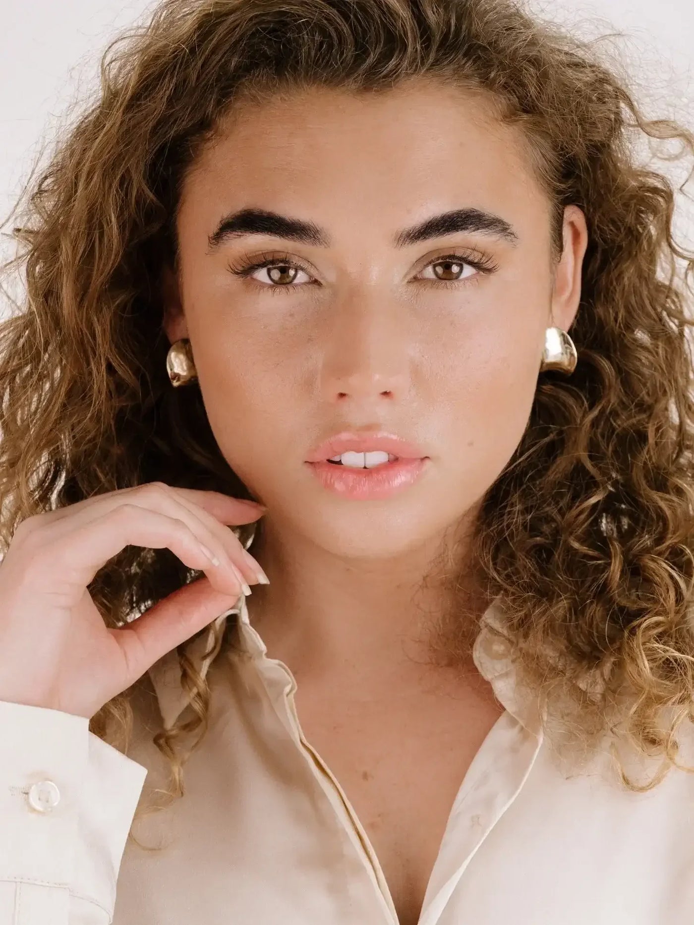 A young woman with curly hair poses, softly touching her face. She wears a light-colored blouse and bold earrings, set against a neutral background, exuding confidence and elegance.