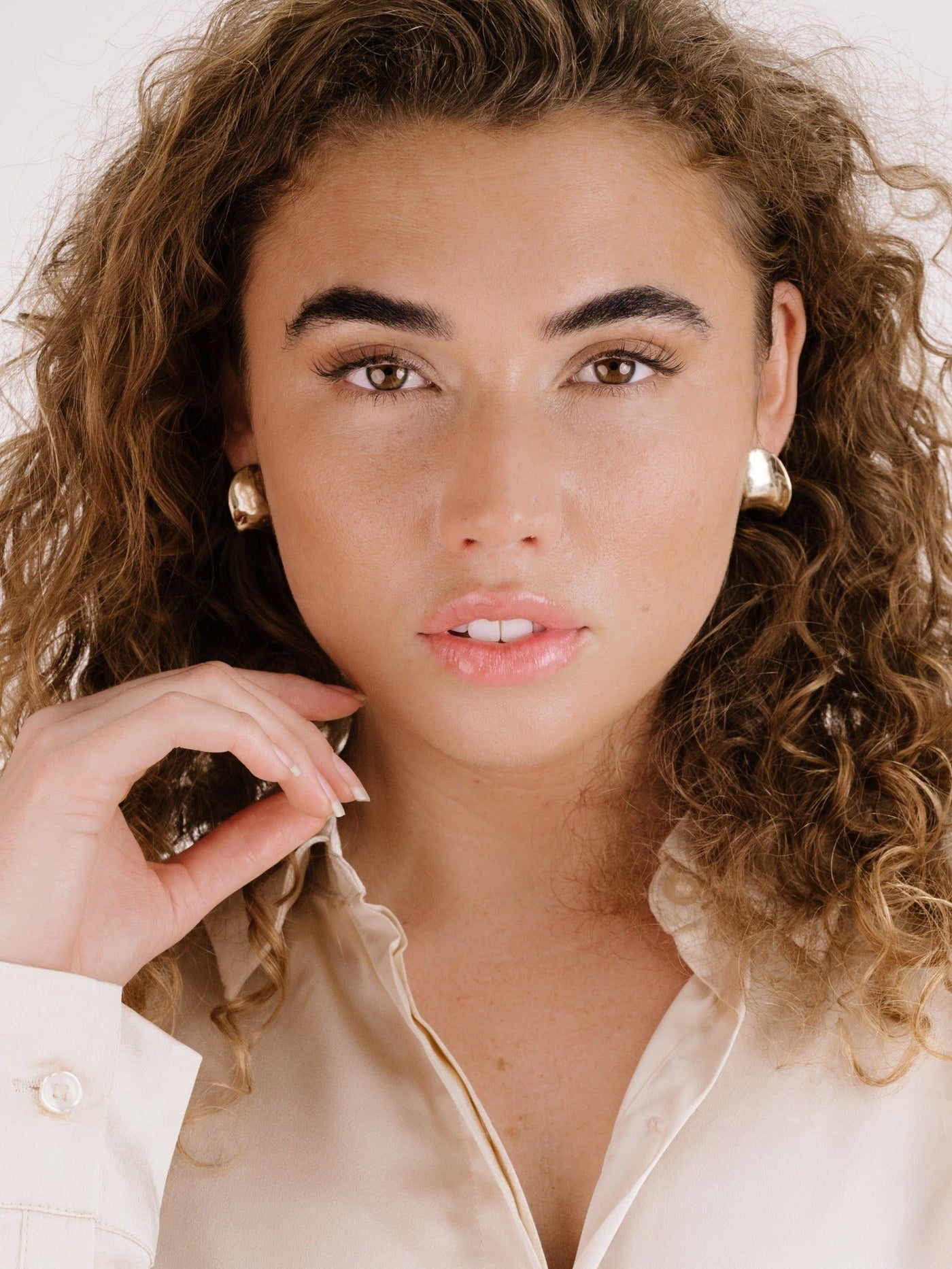 A young woman with curly hair poses, softly touching her face. She wears a light-colored blouse and bold earrings, set against a neutral background, exuding confidence and elegance.