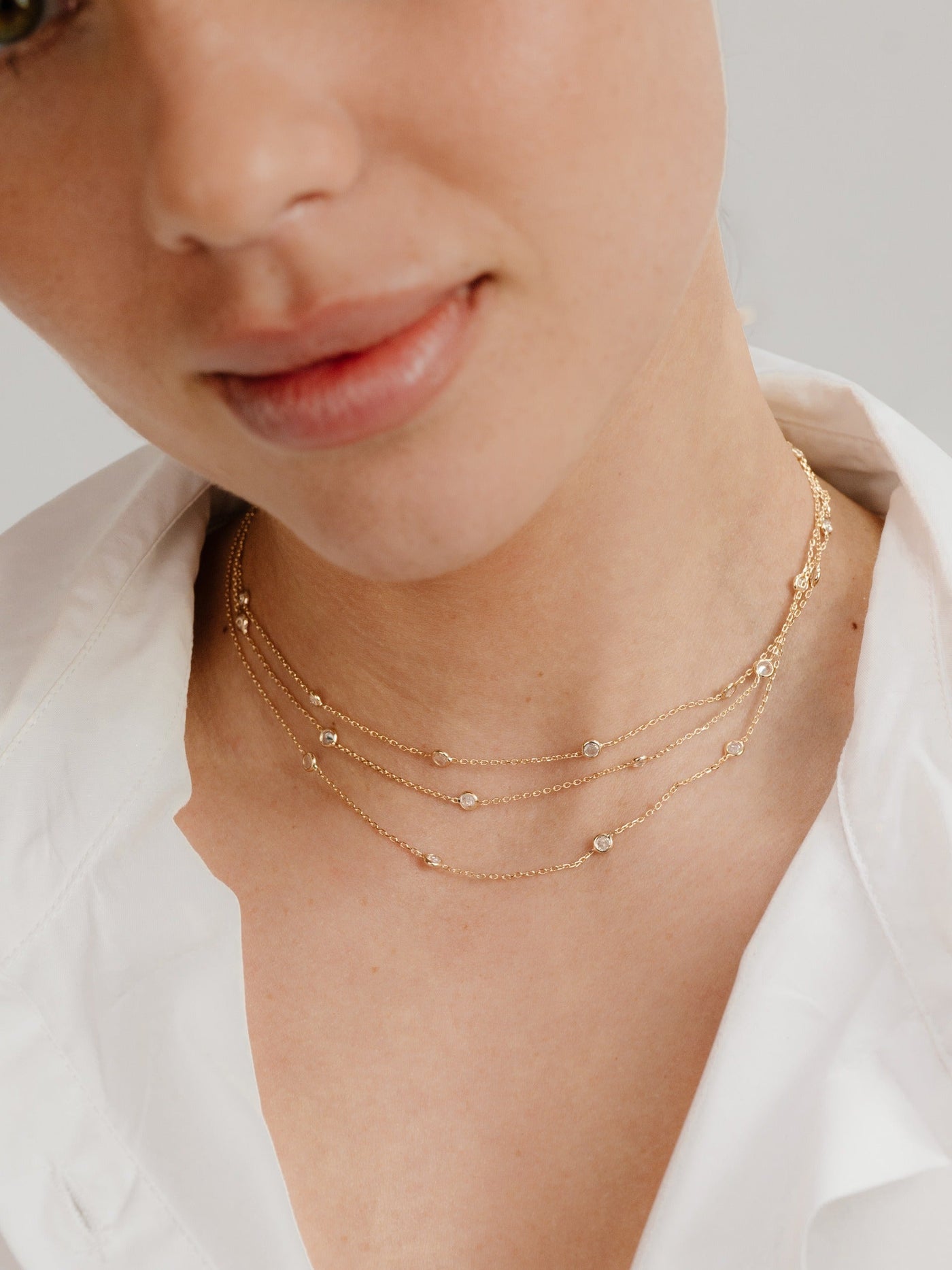 A layered gold necklace with small, sparkling stones adorns the neck of a young woman, who has a soft expression, wearing a white collared shirt against a neutral background.