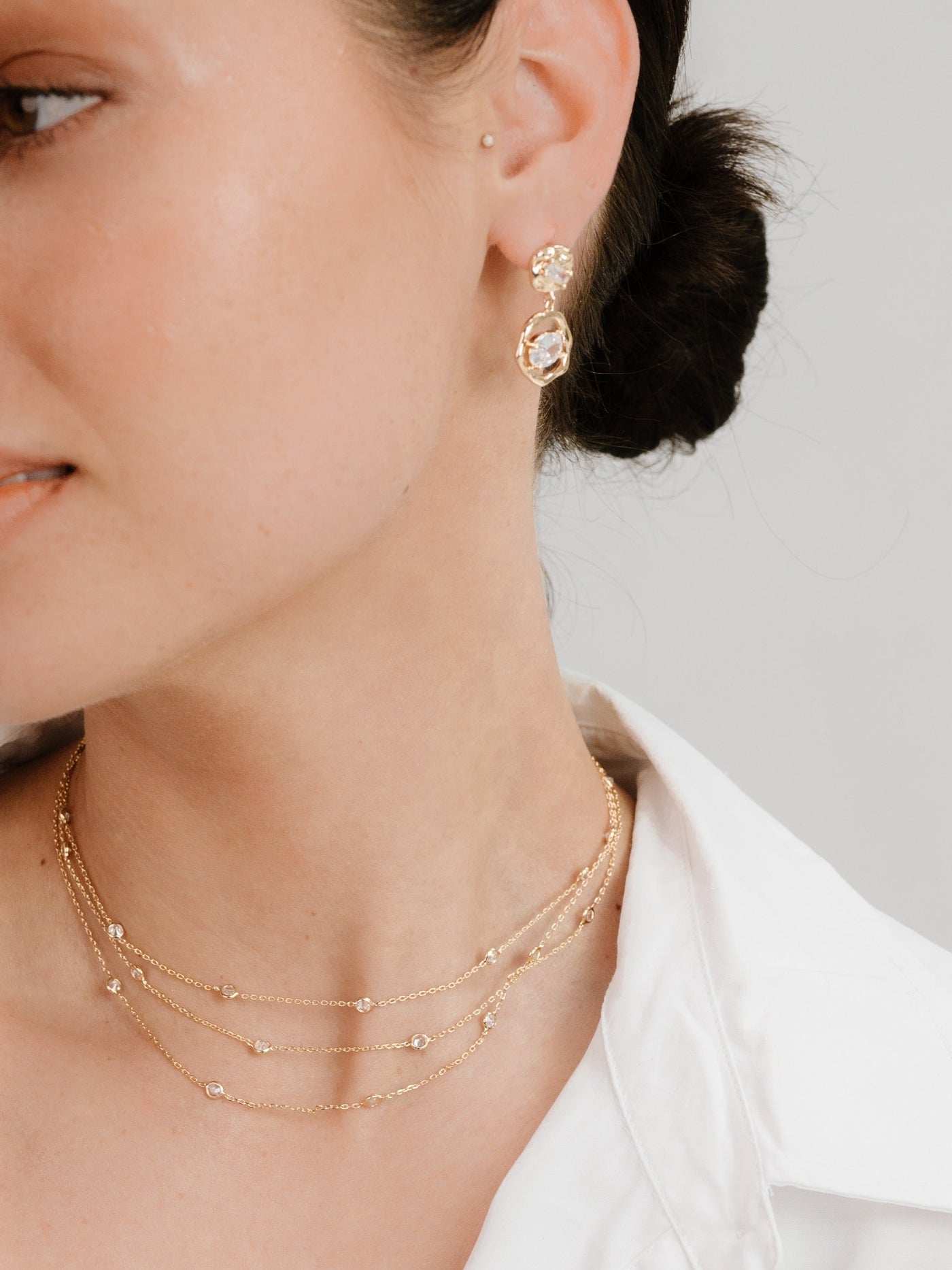 A woman wears elegant gold layered necklaces and decorative earrings, showcasing a refined jewelry aesthetic. She has a neat hairstyle and wears a white blouse, emphasizing her style against a plain background.