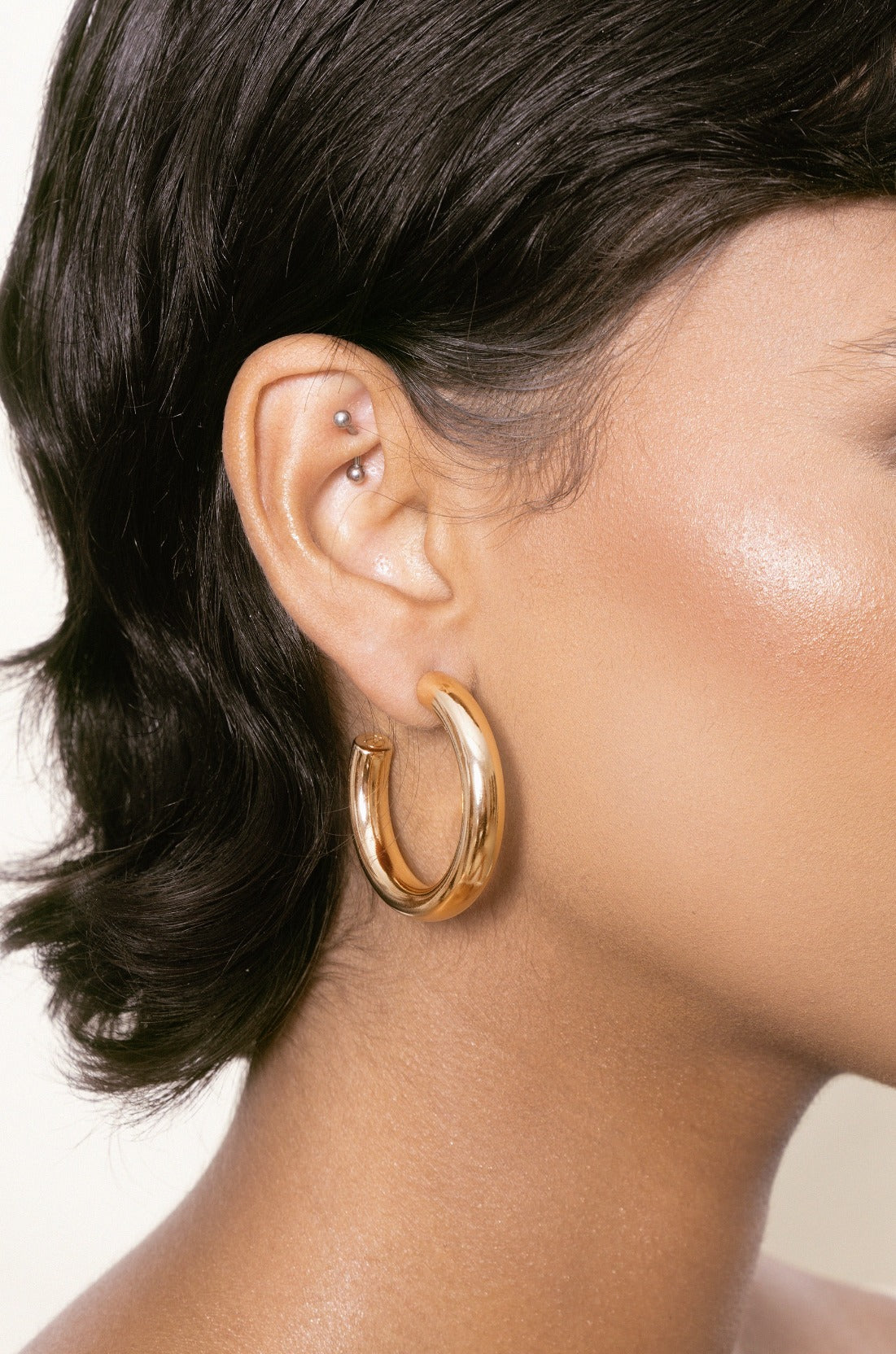 A large, shiny gold hoop earring hangs from a pierced ear, complementing the smooth skin of a person with short, dark hair, set against a light, neutral background.
