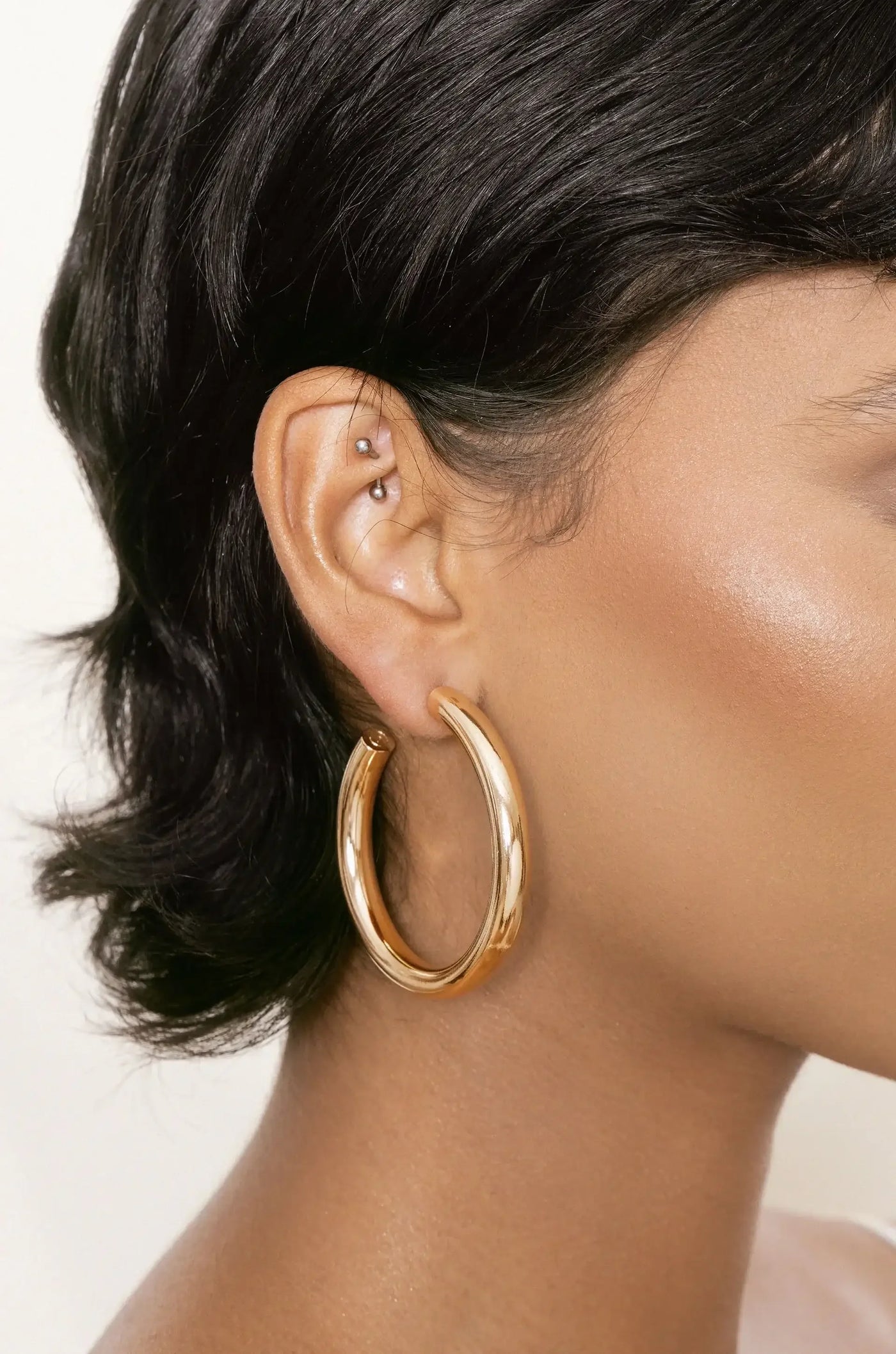 A close-up of a woman's ear features a large, shiny gold hoop earring and a small silver stud piercing, accentuating smooth skin and styled short hair against a neutral background.