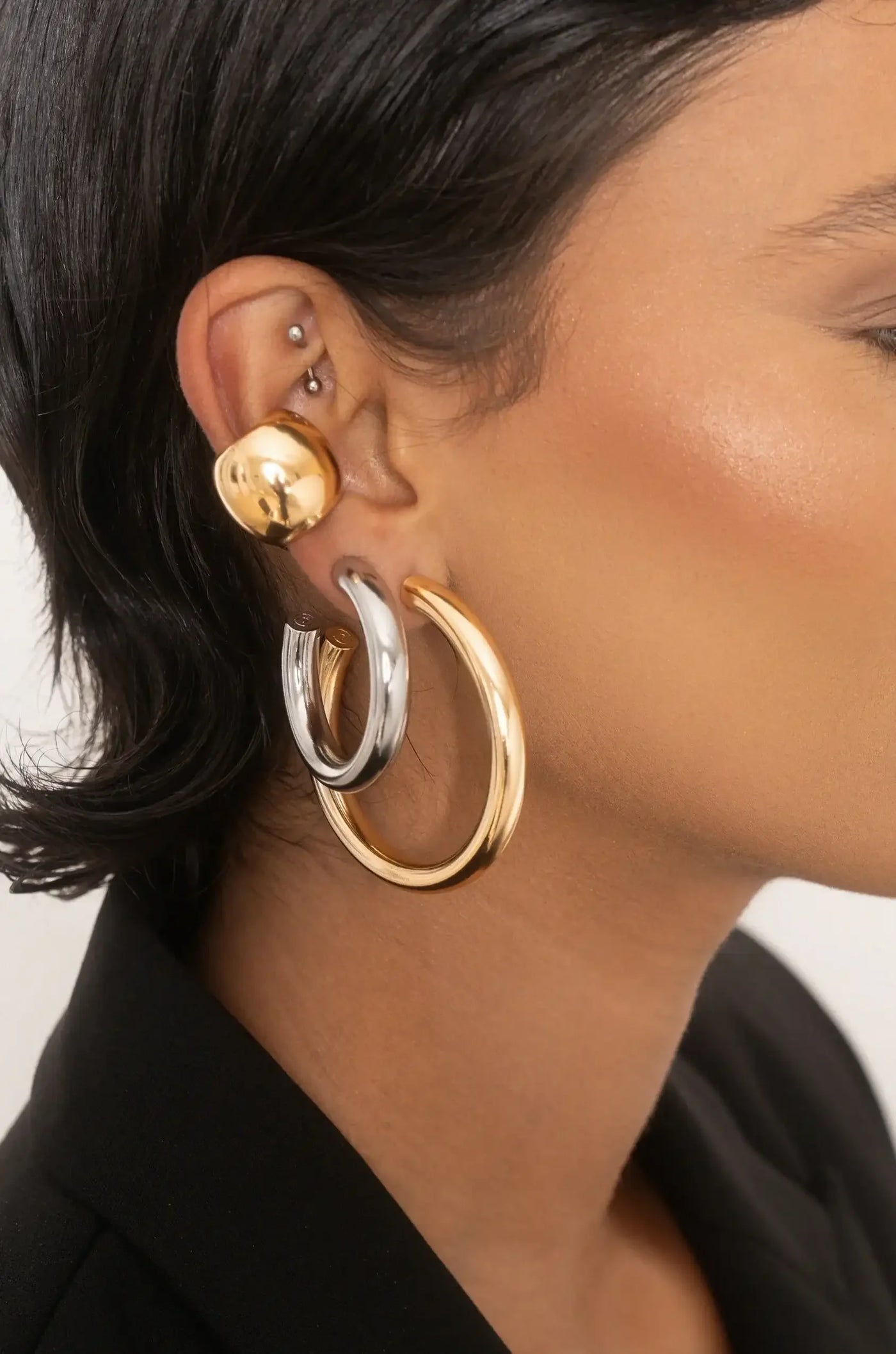 A close-up of a woman's ear showcasing bold earrings. The earrings consist of a shiny gold ball stud, a thick silver hoop, and a large gold hoop. The woman has short black hair and is wearing a dark blazer.