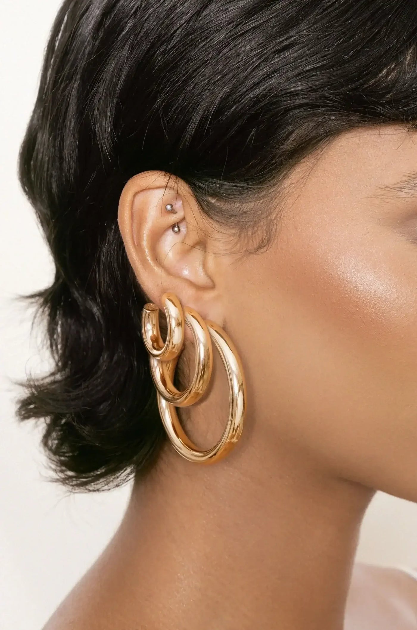 A close-up of an ear showcases multiple shiny, gold hoop earrings layered together. The ear features a small metallic stud, set against a smooth, neutral-toned background.