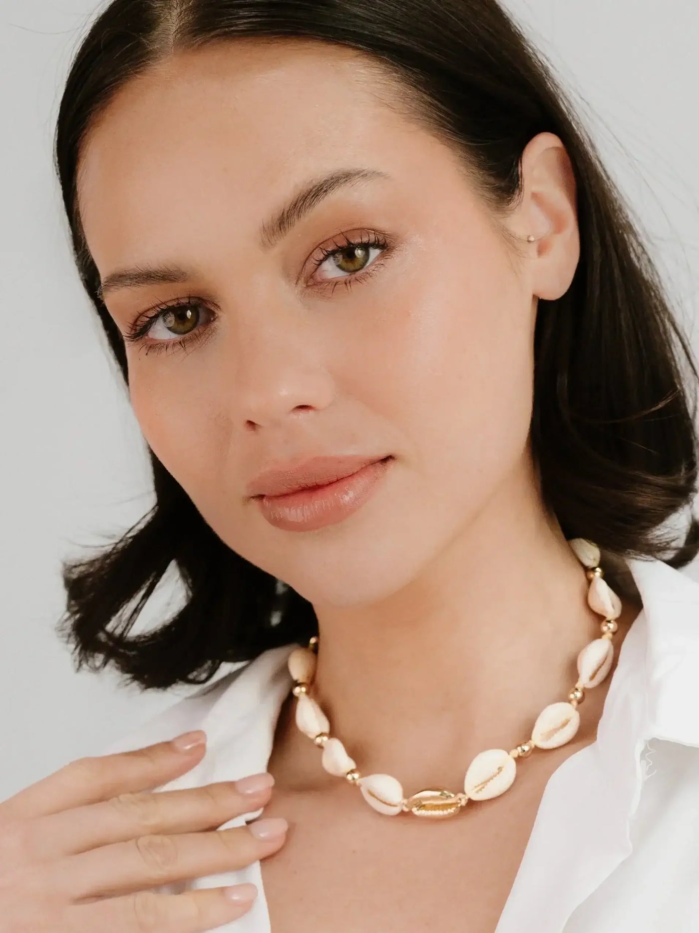 A woman with shoulder-length dark hair gently touches a shell necklace around her neck, smiling softly. She wears a white shirt, and the background is a light, neutral color.