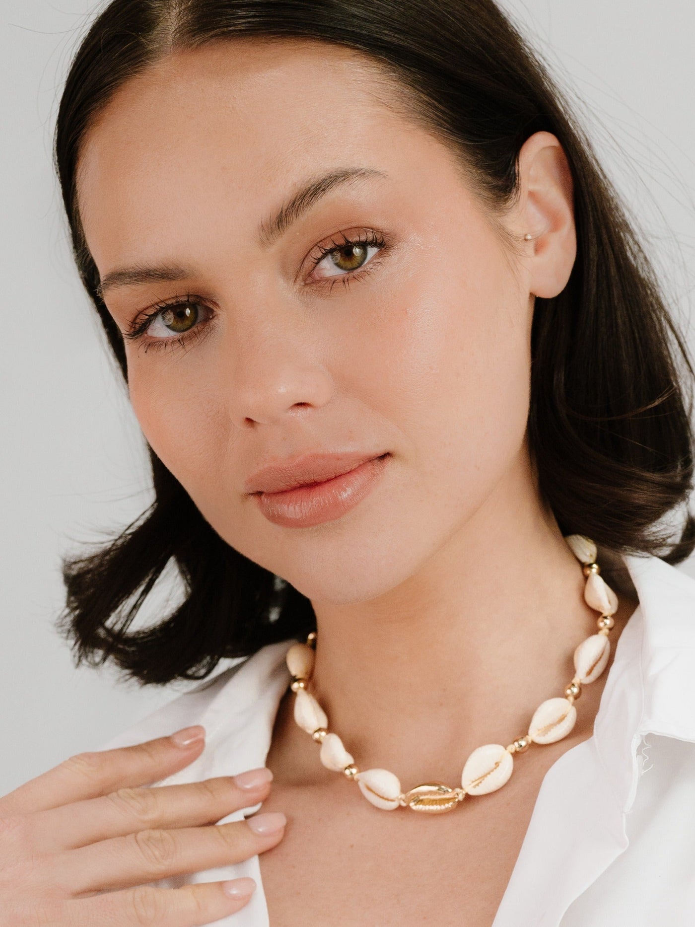 A woman with shoulder-length dark hair gently touches a shell necklace around her neck, smiling softly. She wears a white shirt, and the background is a light, neutral color.