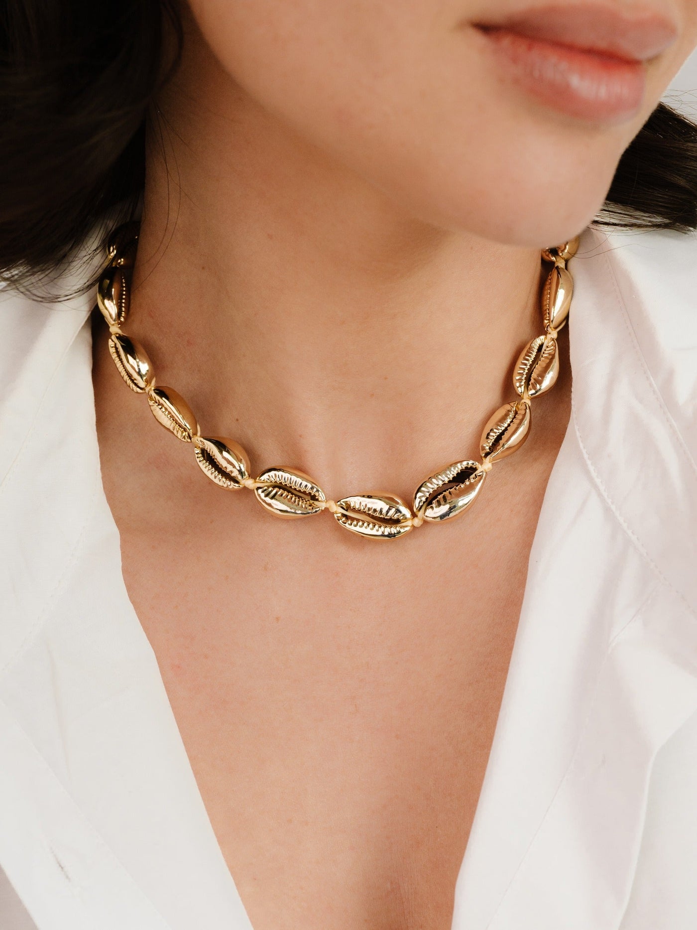 A gold statement necklace, featuring oval links with intricate designs, adorns the neck of a woman in a white blouse. The setting is softly lit, highlighting the jewelry's shine.