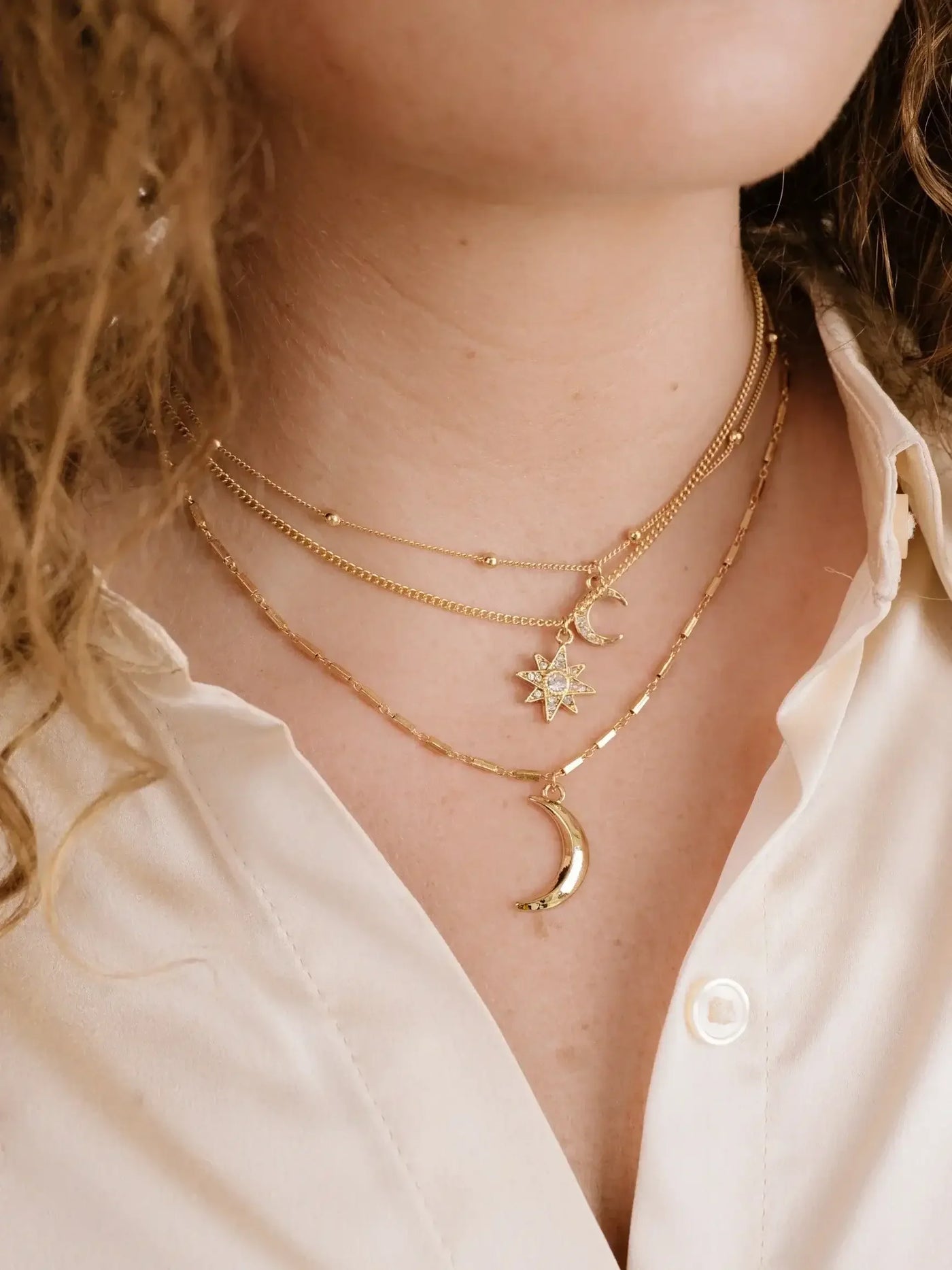 Layered gold necklaces adorn a woman's neck, featuring a crescent moon and a star pendant. She wears a light-colored shirt with an open collar, in a softly lit environment.
