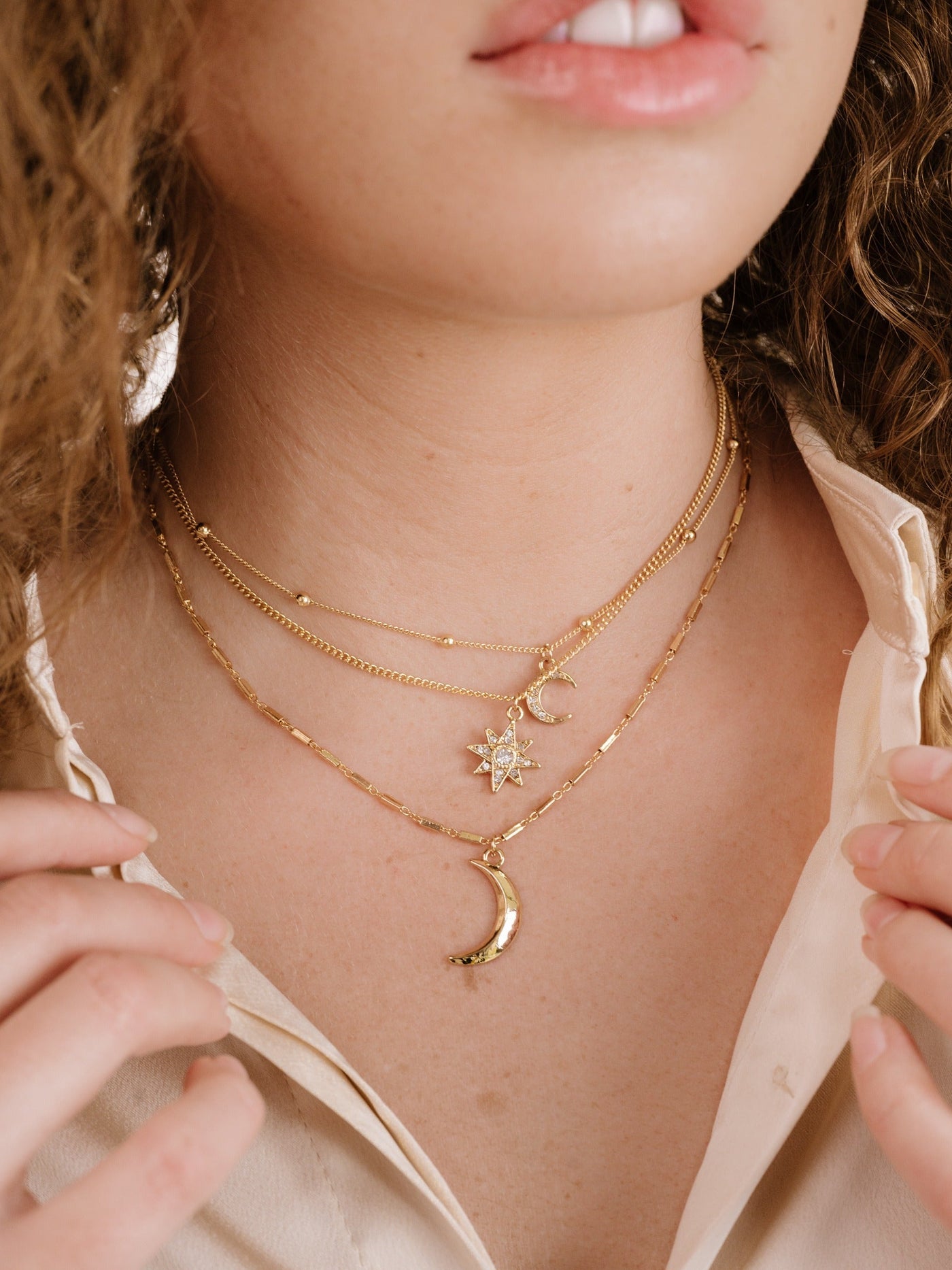 A person’s neck adorned with layered gold necklaces featuring a crescent moon, a star pendant, and delicate chains. They gently touch their collar, wearing a light, open shirt.