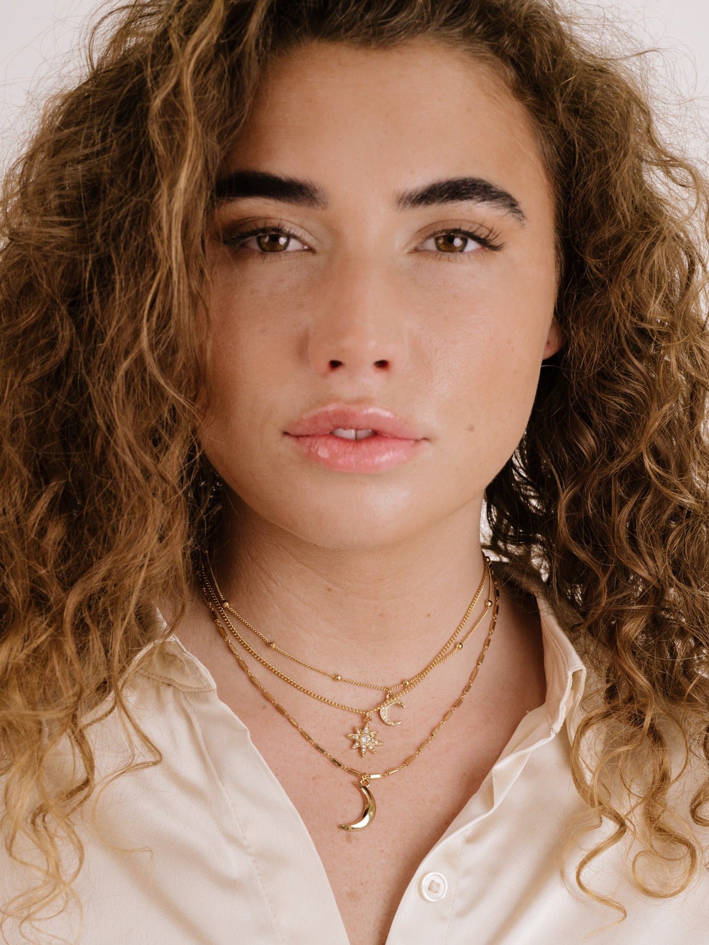 A young woman with curly hair gazes directly at the viewer. She wears a light-colored blouse and layered gold necklaces featuring moon and star charms, set against a plain background.