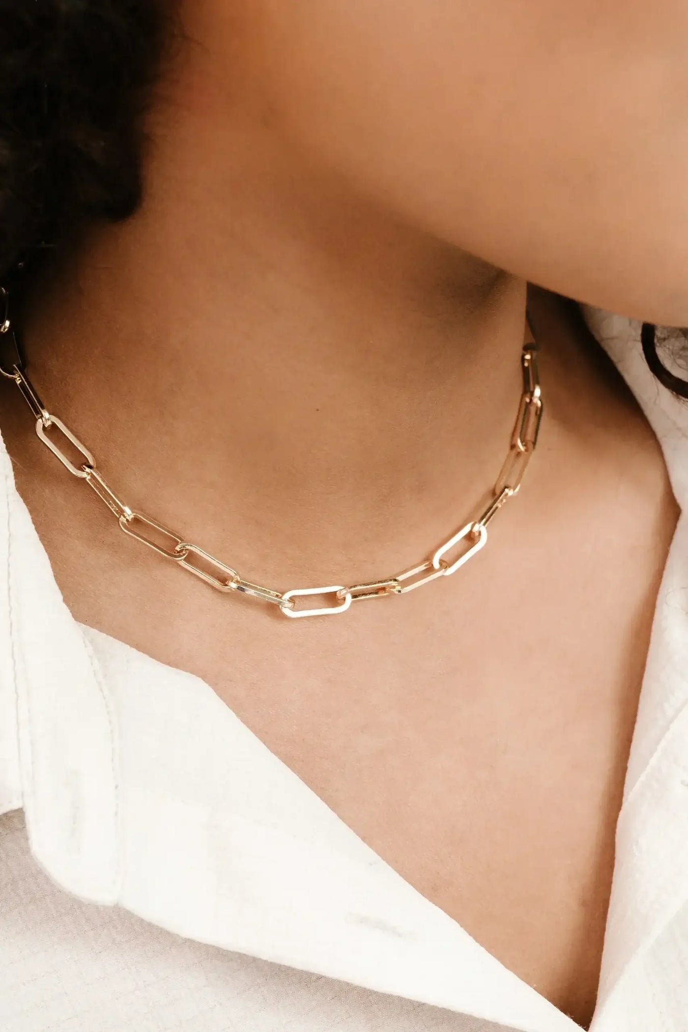 A gold chain necklace with elongated links rests on a model's neck, complementing a white collared shirt. The setting appears casual, focusing on the jewelry's elegant design.