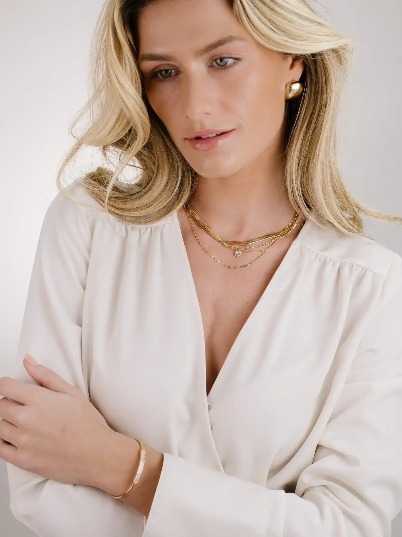 A woman with long, wavy blonde hair wears layered gold necklaces and an elegant blouse, gently resting her arms and looking thoughtfully off-camera against a soft, neutral background.
