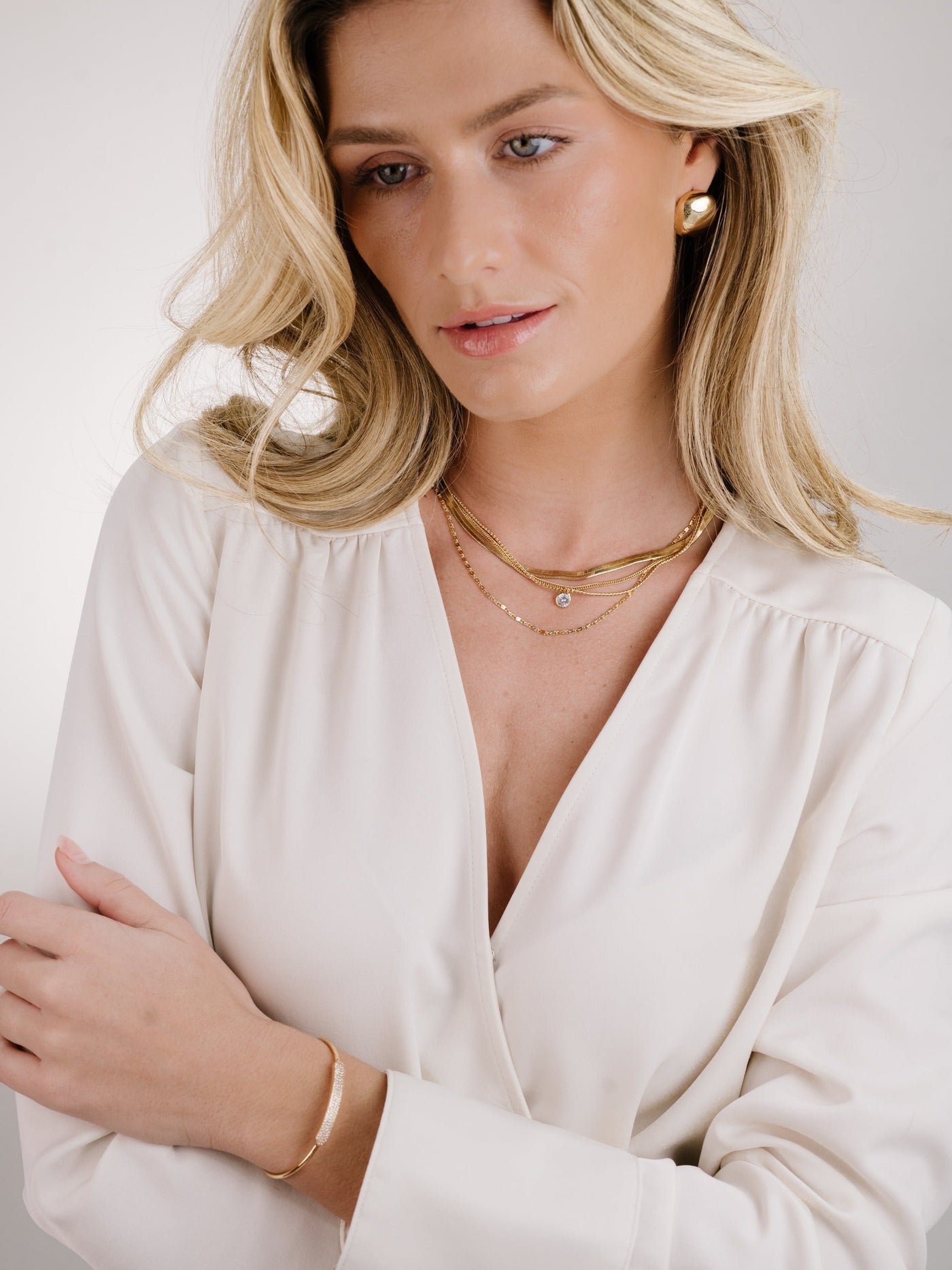 A woman with long, wavy blonde hair wears layered gold necklaces and an elegant blouse, gently resting her arms and looking thoughtfully off-camera against a soft, neutral background.