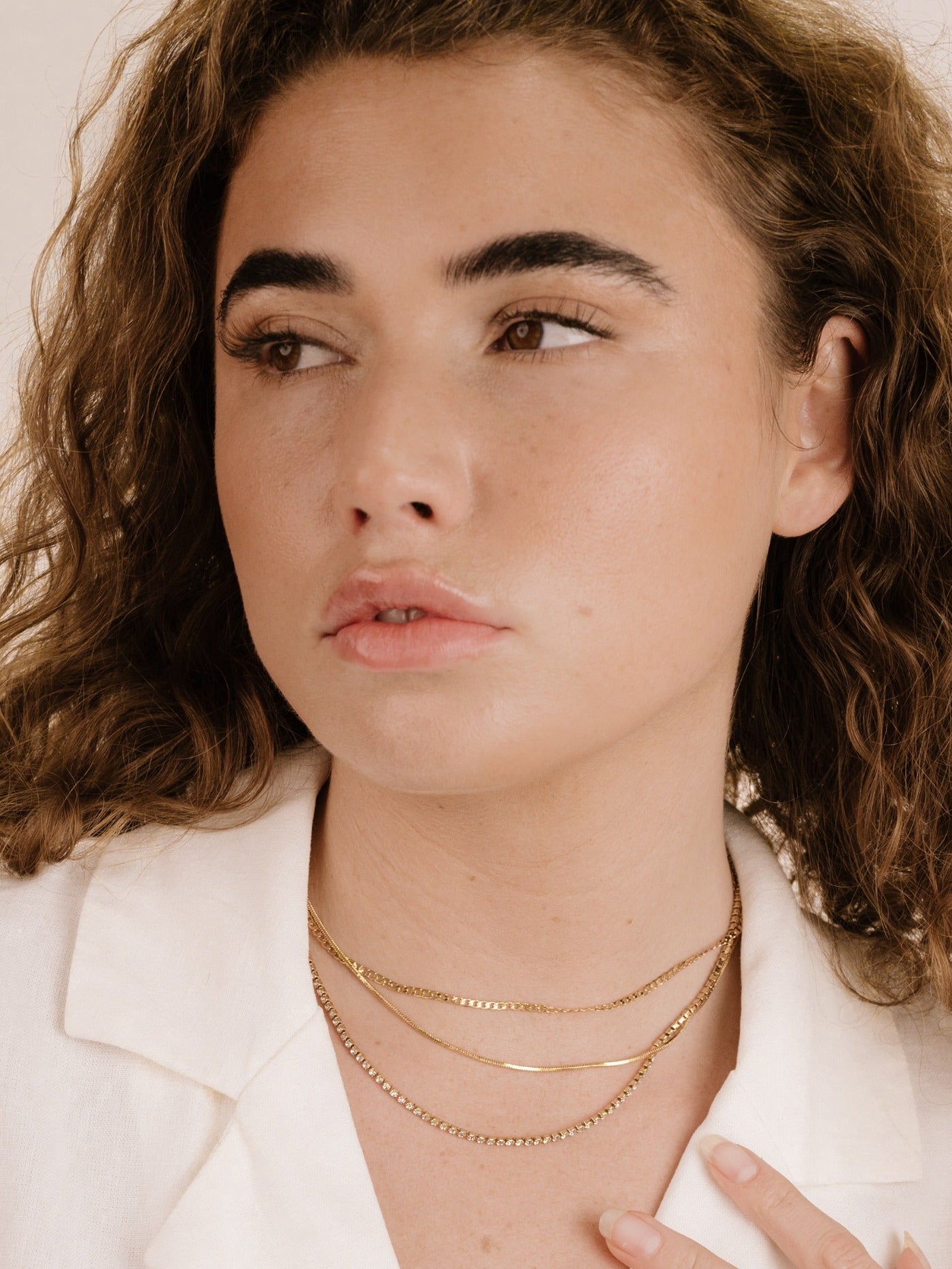 A person with medium-length curly hair gazes thoughtfully, wearing layered gold necklaces. Their skin has a natural glow, and they are dressed in a light-colored collared shirt against a neutral background.