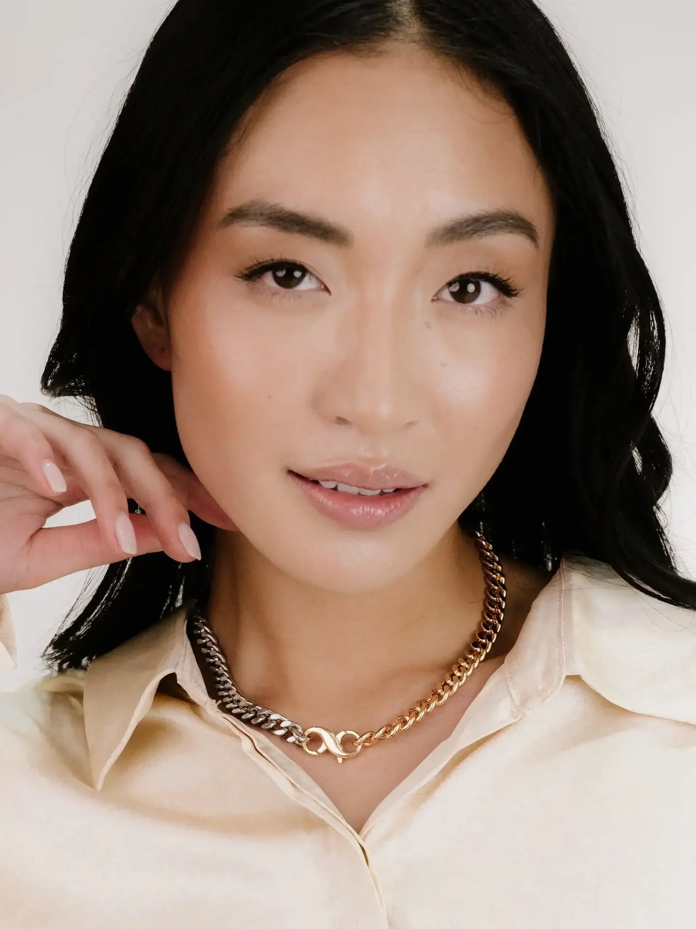 A woman with long black hair poses with a hand gently touching her neck, showcasing a mixed metal chain necklace against a neutral background, wearing a light-colored button-up shirt.