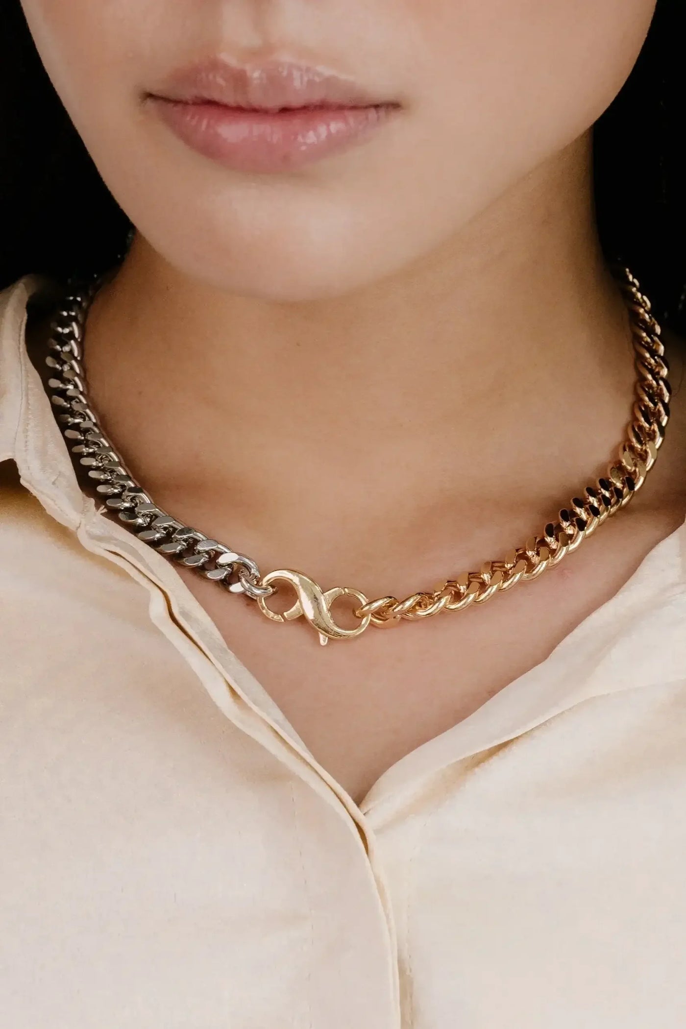 A silver and gold chain necklace with an infinity symbol is worn around the neck of a person, who has smooth lips and is dressed in a light-colored, button-up shirt.